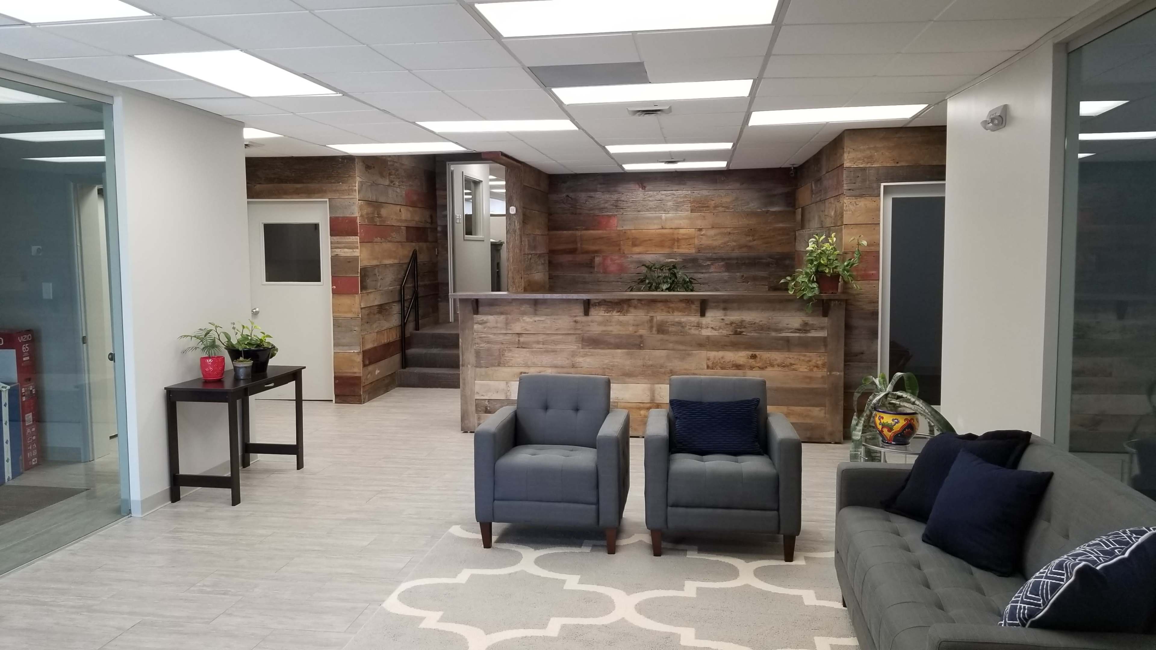A modern office reception area with a wooden front desk, two gray armchairs, and a gray couch, surrounded by plants and a patterned rug.