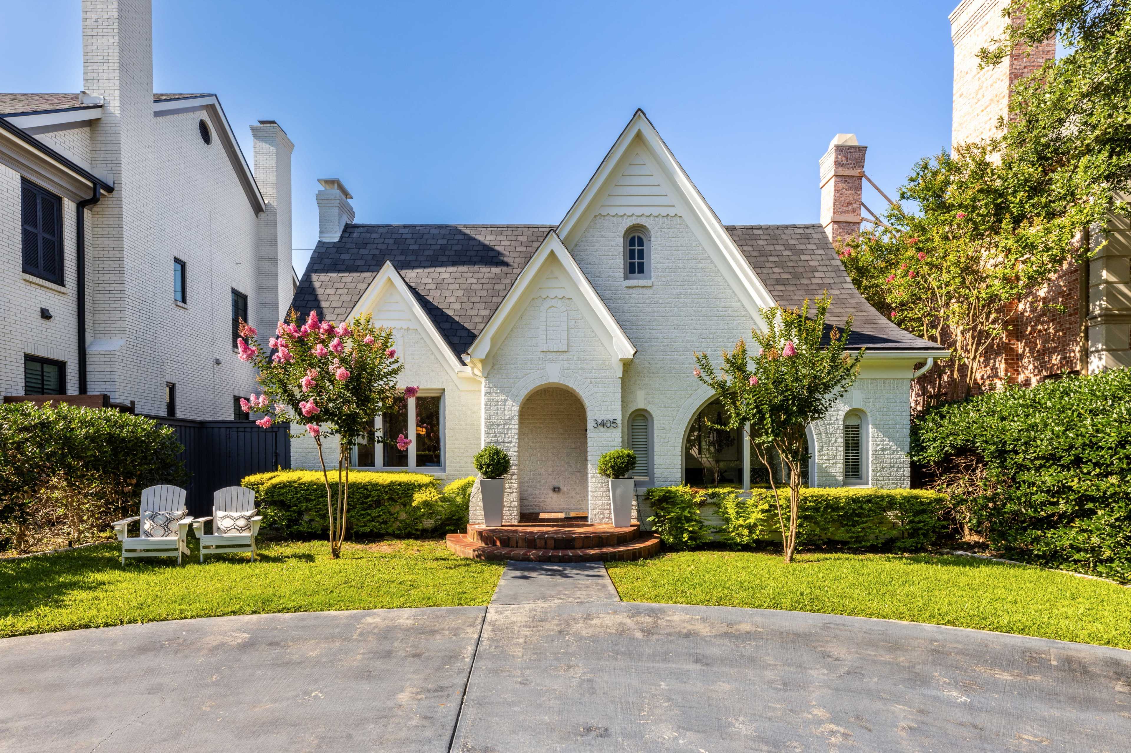 The image shows a white, single-family house with a steeply pitched roof, flanked by manicured lawns and two adirondack chairs in the front yard.