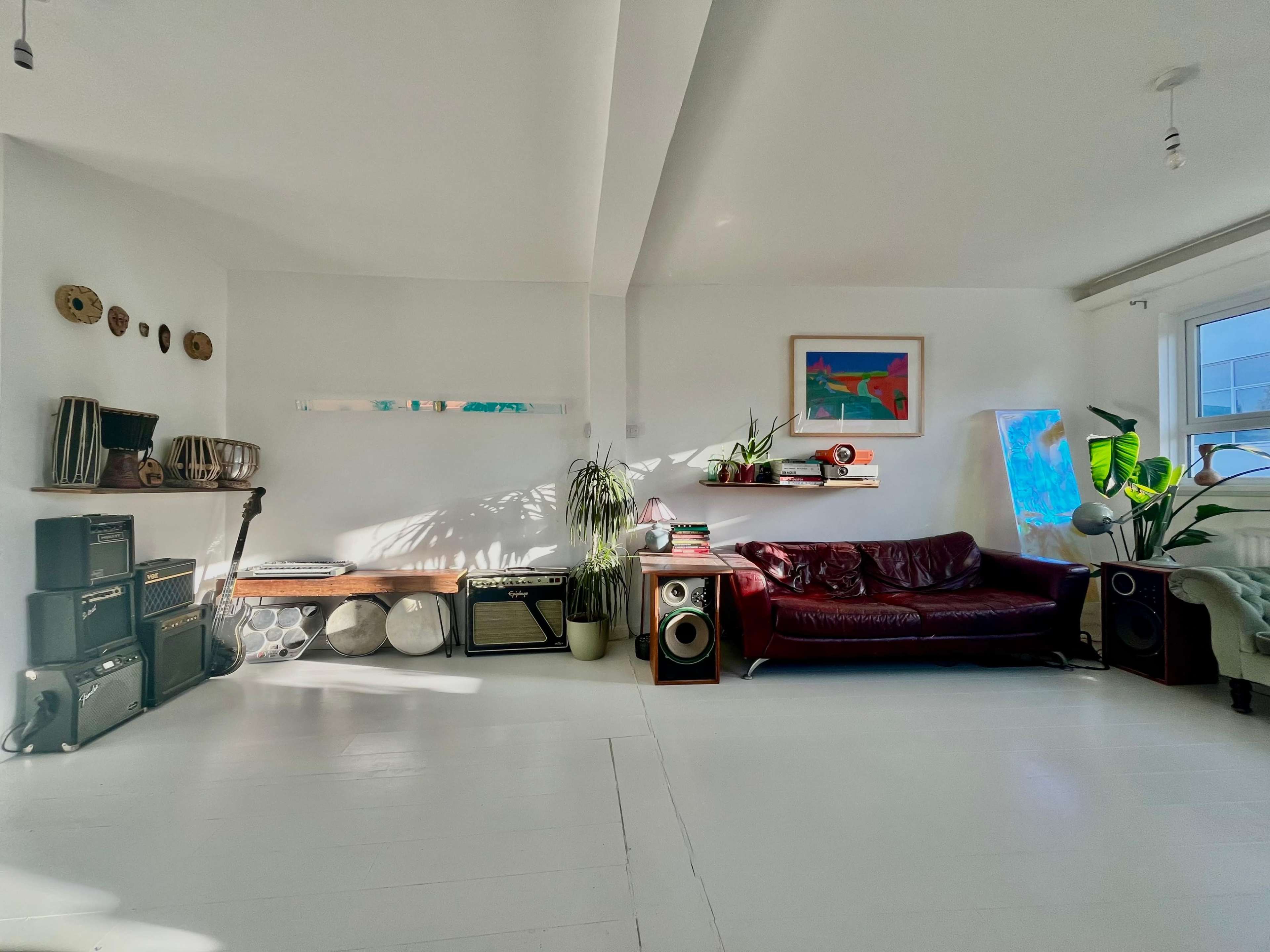 The image shows a bright, modern living room featuring a burgundy couch, various plants, and shelving with musical instruments and artwork.