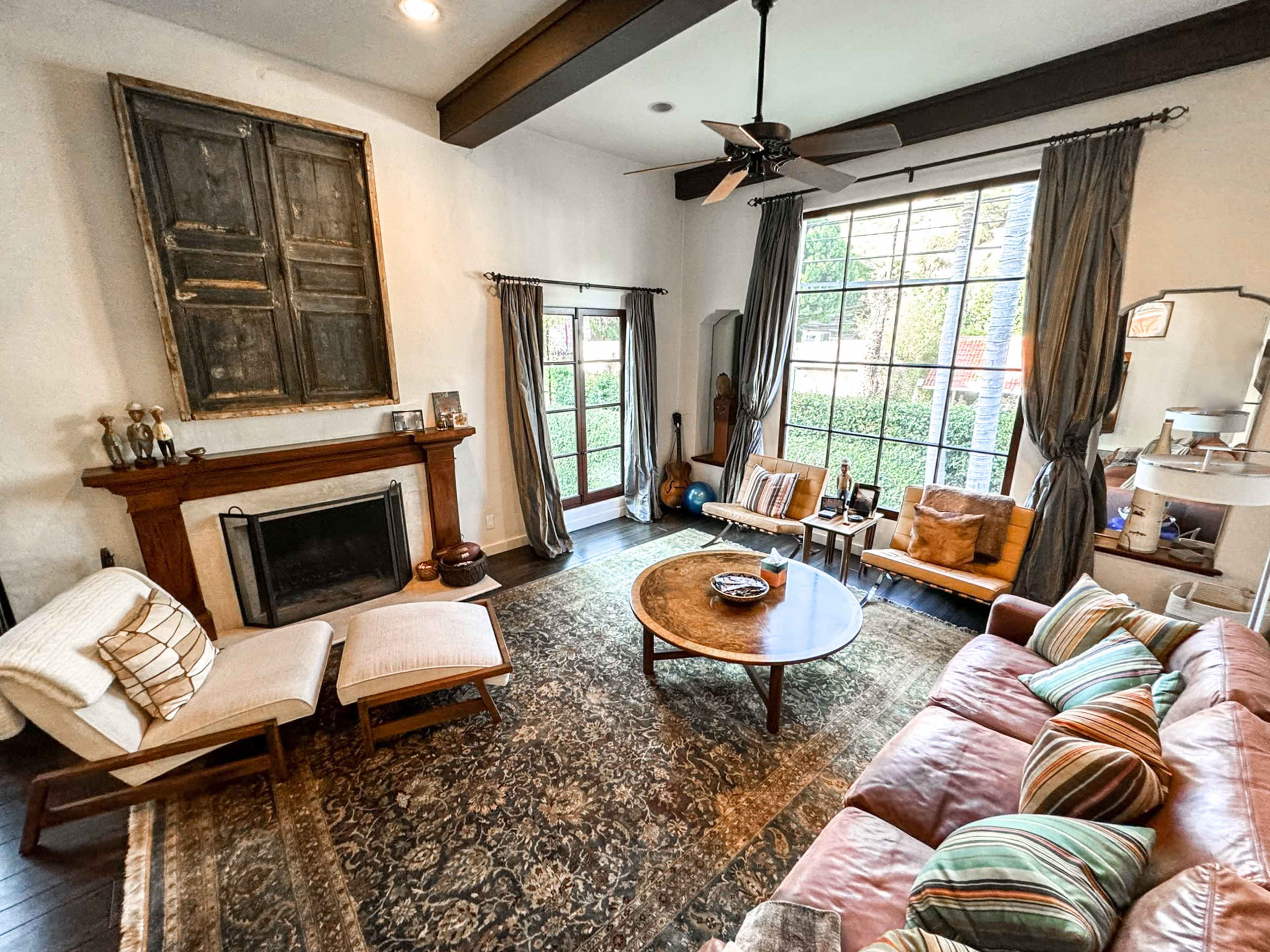 The living room features a large window, a fireplace with a wooden mantel, two lounge chairs, a round coffee table, and a sofa with patterned cushions.