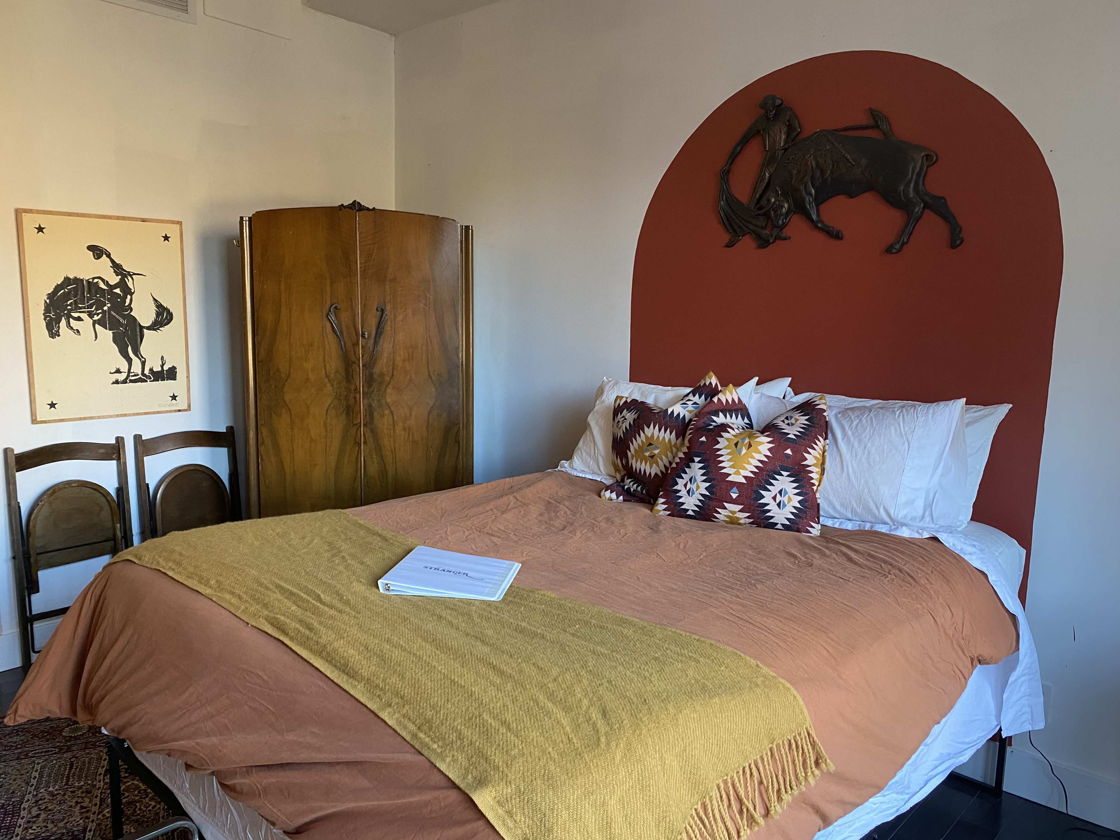 A cozy bedroom featuring a large bed with a brown spread and patterned pillows, a wooden wardrobe, a framed cowboy-themed artwork on the wall, and a distinctive red arch with a bull sculpture above the headboard.