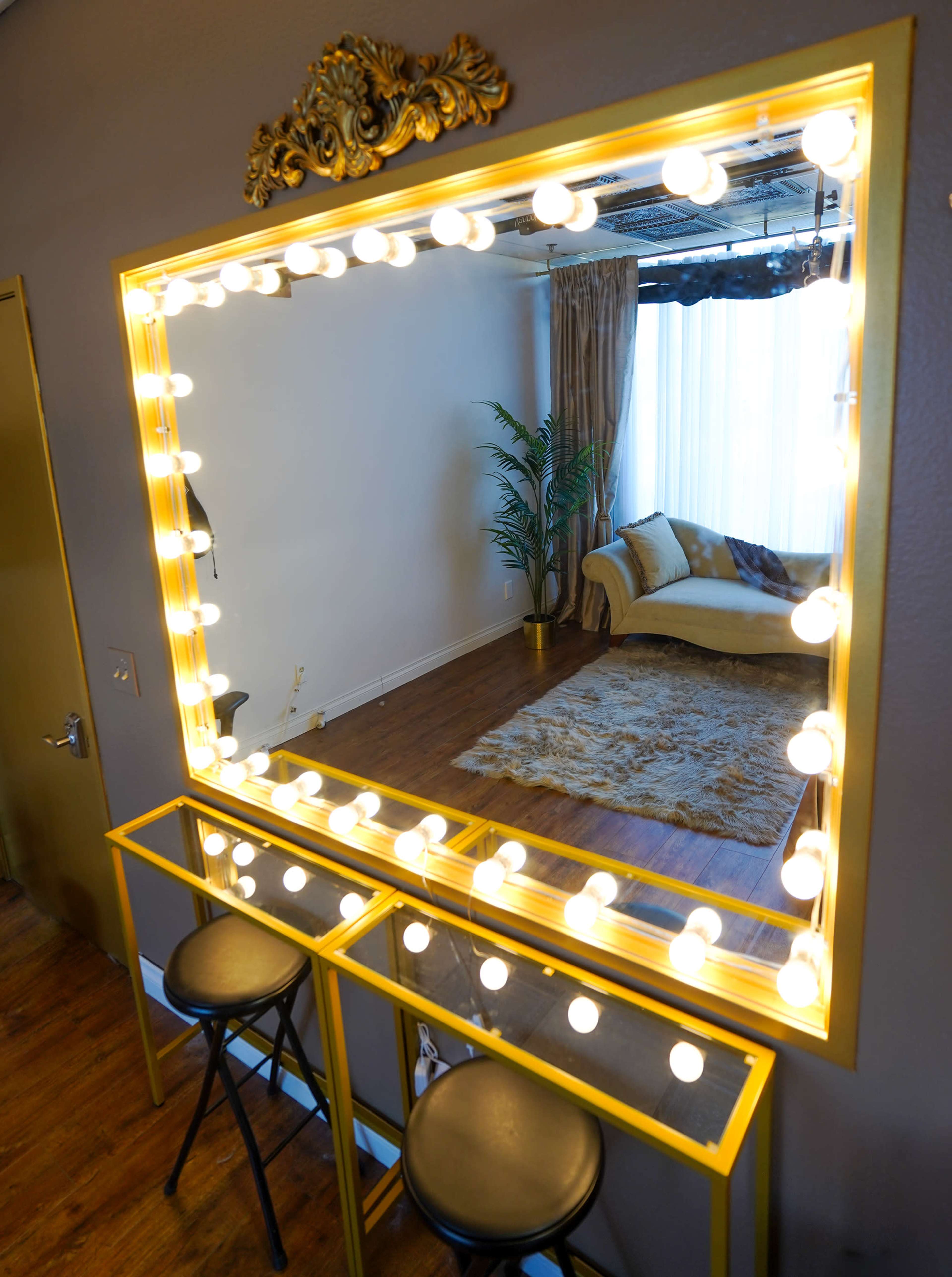 A large illuminated vanity mirror with a decorative frame is positioned above a narrow table with two stools, reflecting a room with a couch and a decorative plant in the background.