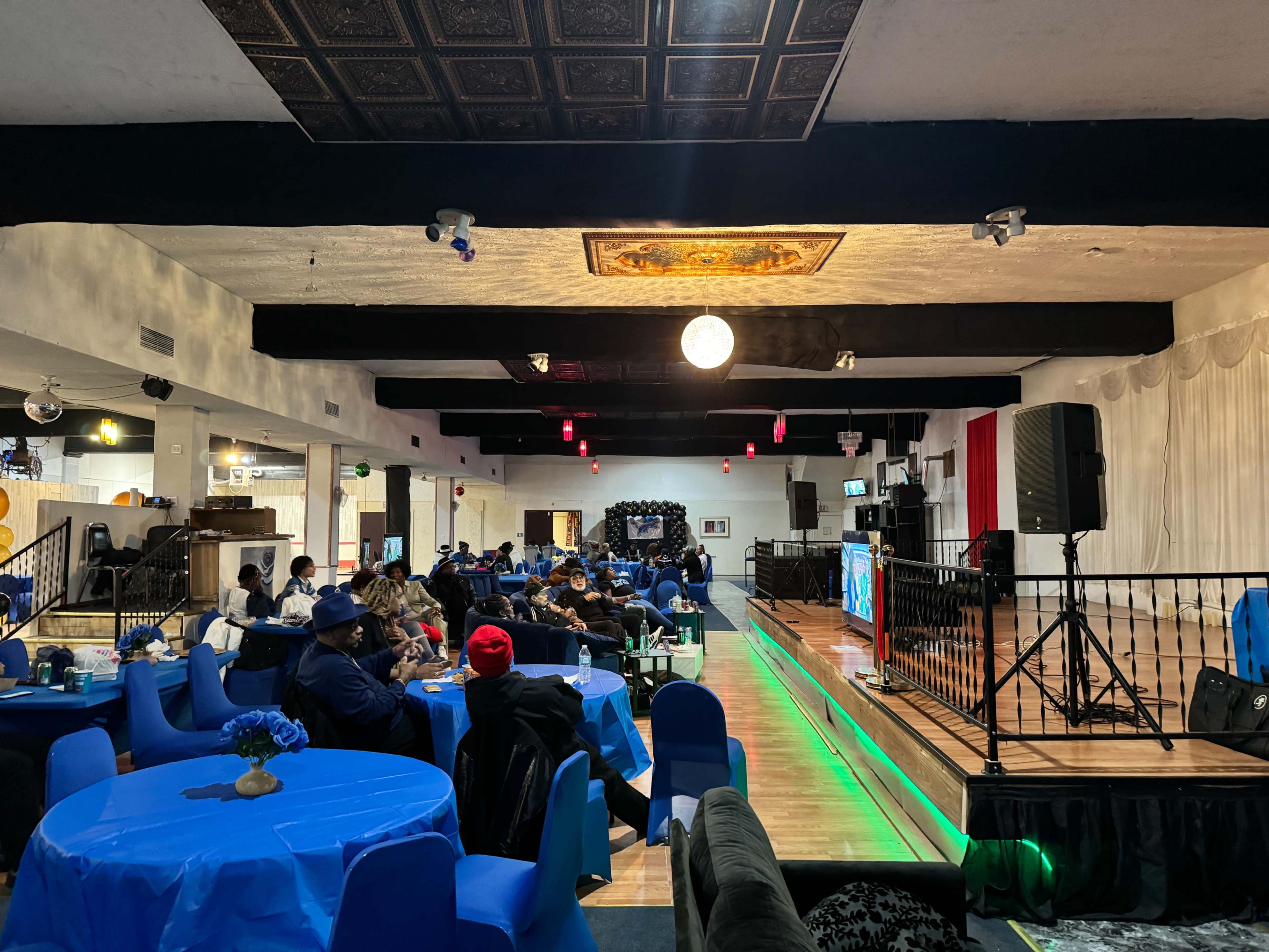 The image shows a spacious indoor venue with blue tables and chairs, where a group of people are seated, facing a stage with a microphone and a screen.