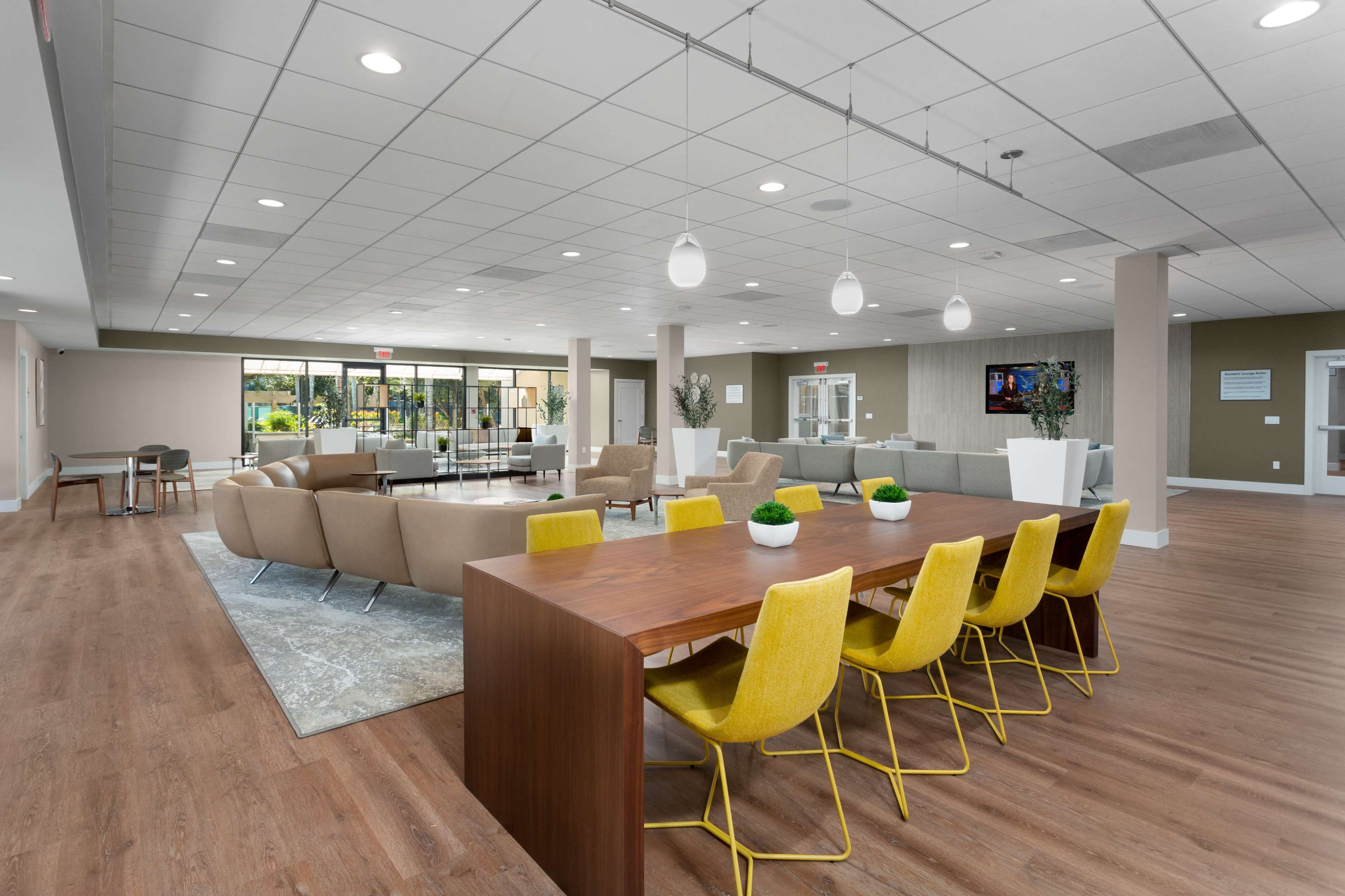 A modern interior space featuring a dining area with a long wooden table and yellow chairs, along with a lounge area that includes beige sofas, all under bright lighting.
