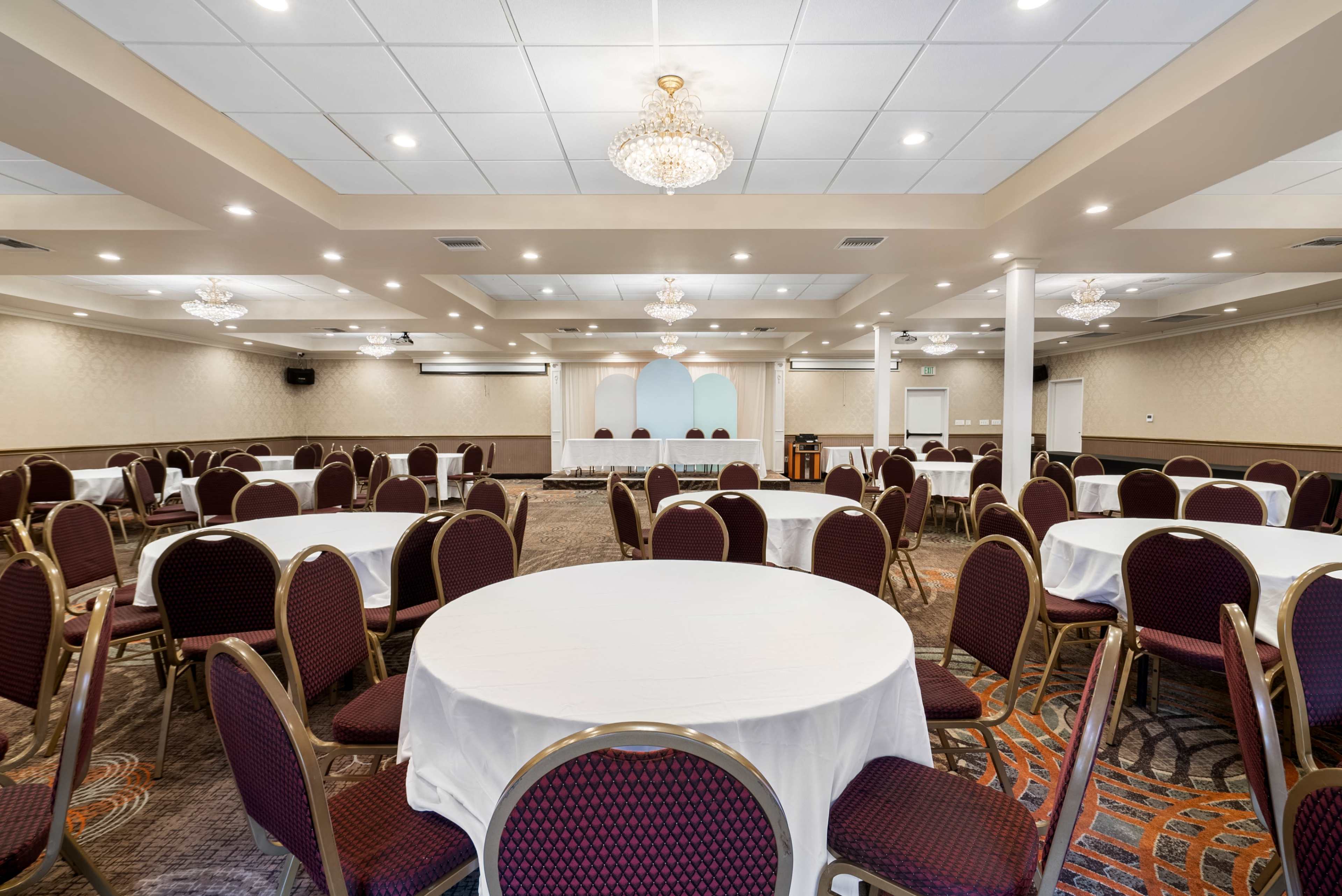 A spacious banquet hall features round tables with white tablecloths and elegant chandeliers overhead.