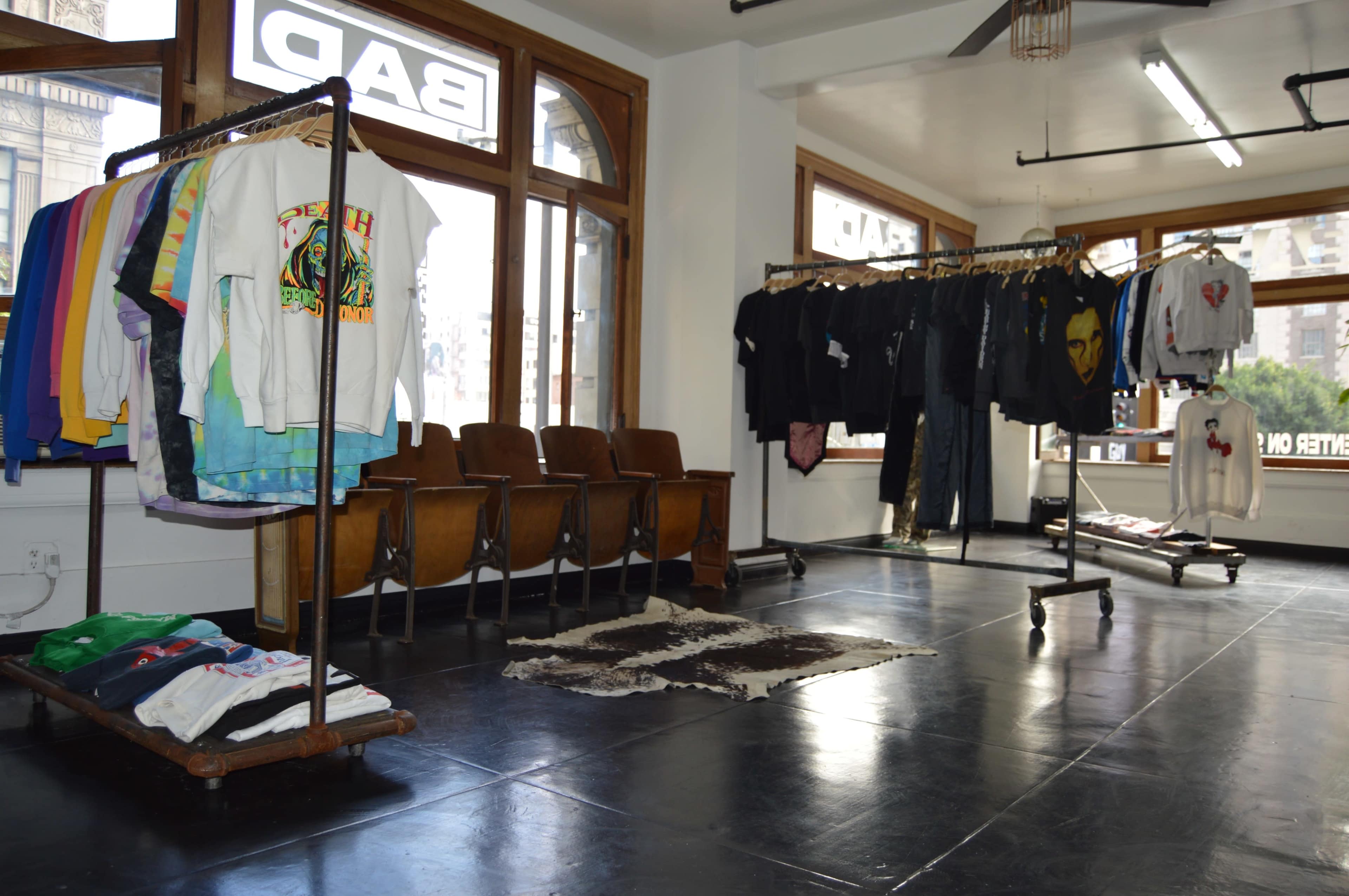 The image shows a clothing store interior with two clothing racks displaying various garments, and a row of wooden chairs in front of large windows.