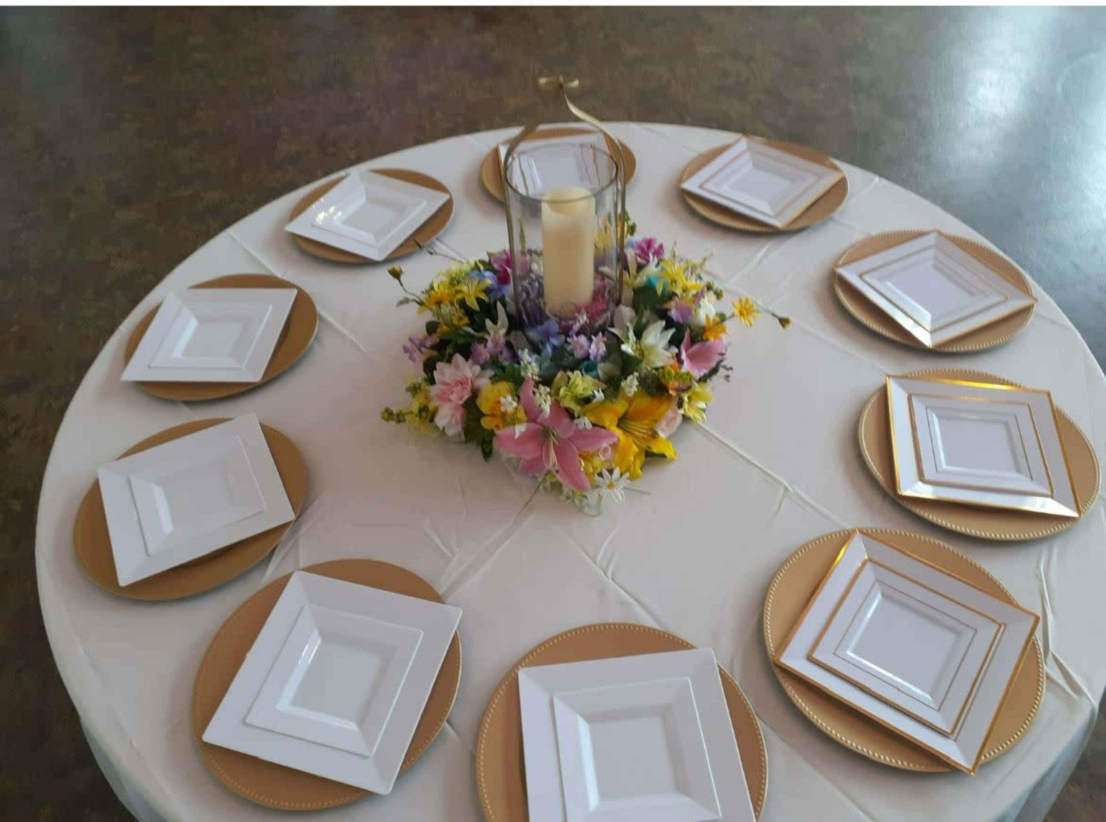 A round table is set with ten white square plates arranged in a circular pattern around a floral centerpiece with a candle.