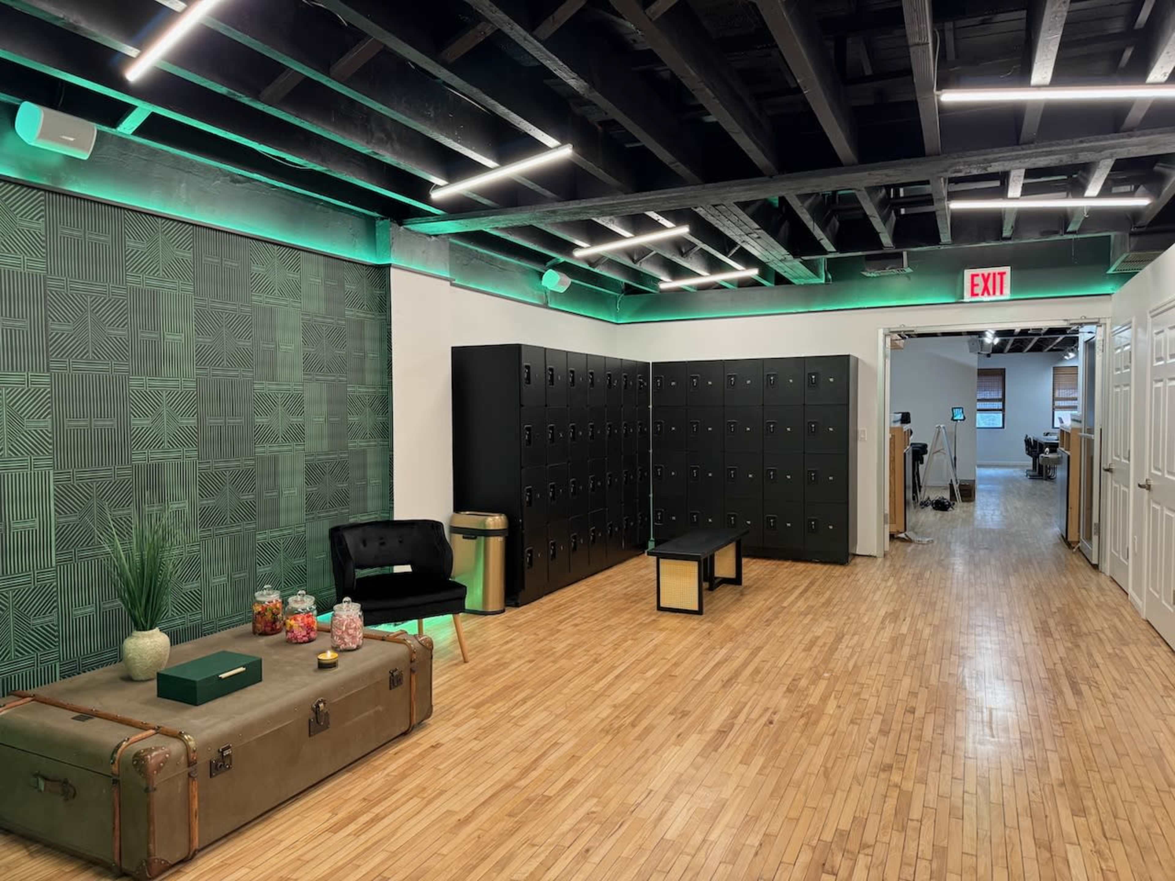The image shows a modern hallway featuring wooden flooring, green accent lighting, black lockers on one side, a dark sofa, and a decorative trunk with plants and candy jars.