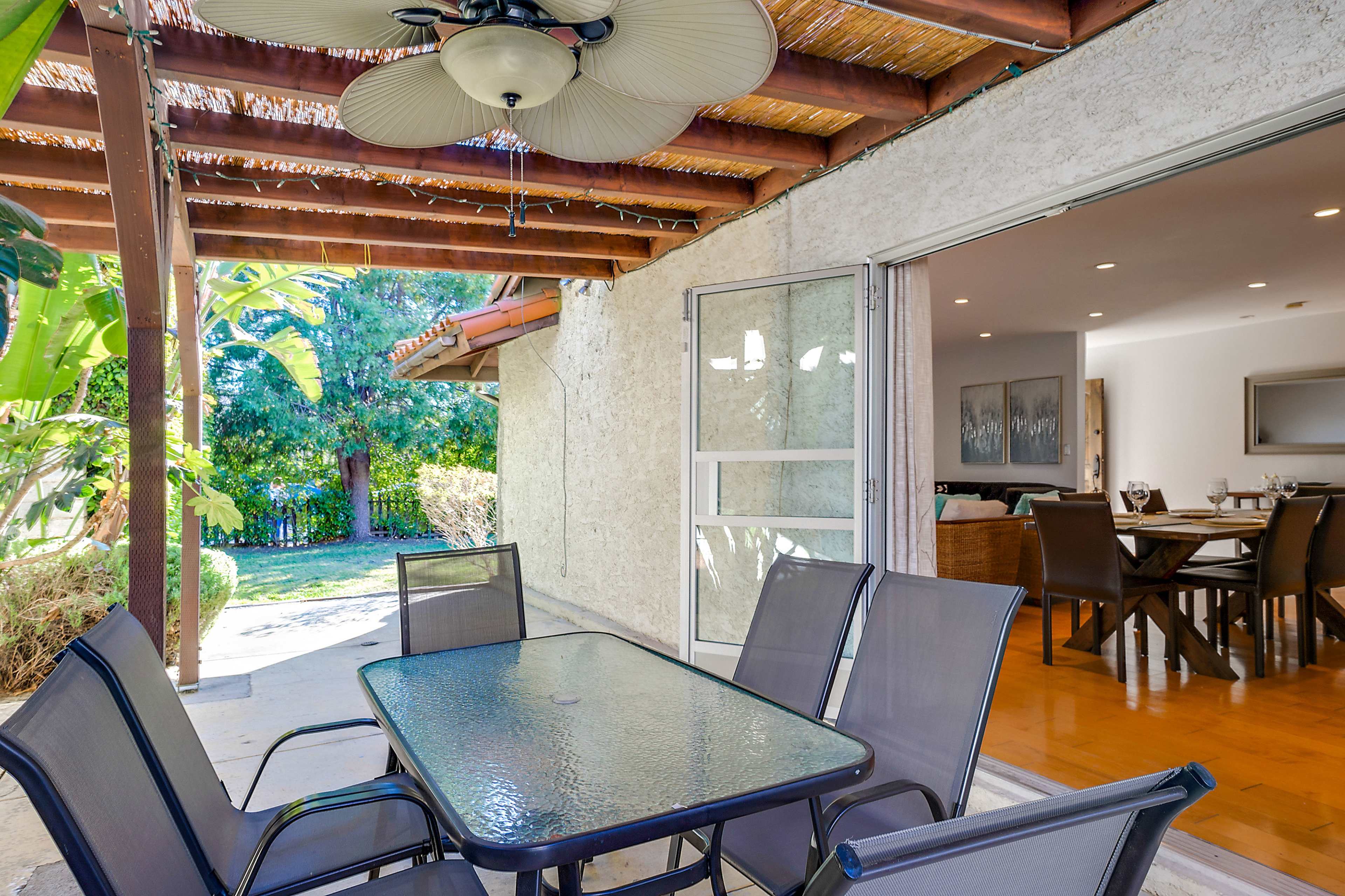 A patio area features a glass-top table surrounded by black chairs, under a thatched roof with a ceiling fan, adjacent to a yard with green foliage.