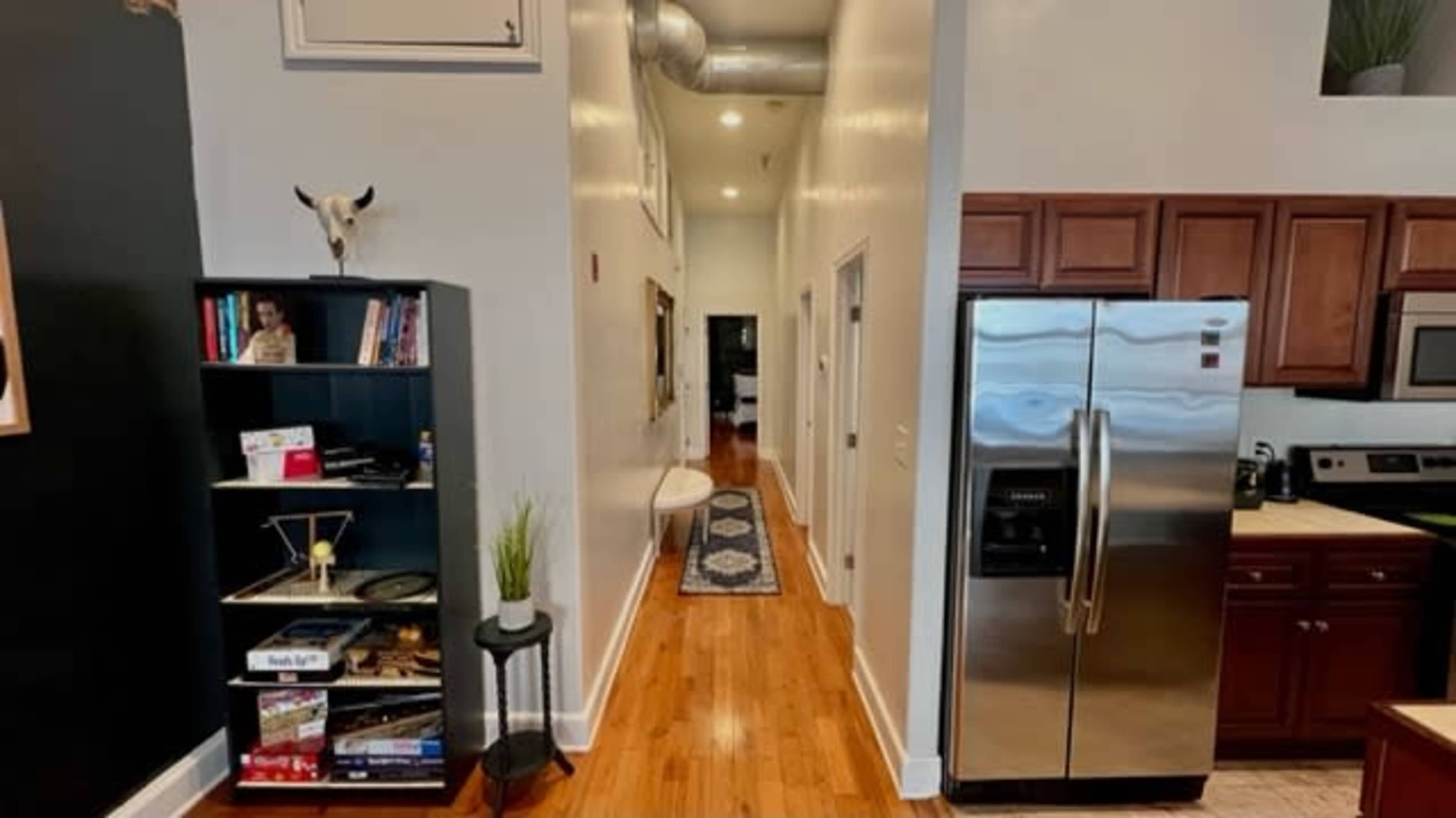 A narrow hallway leading into an apartment, with a bookshelf on the left, a white bench along the wall, and a kitchen area visible to the right.