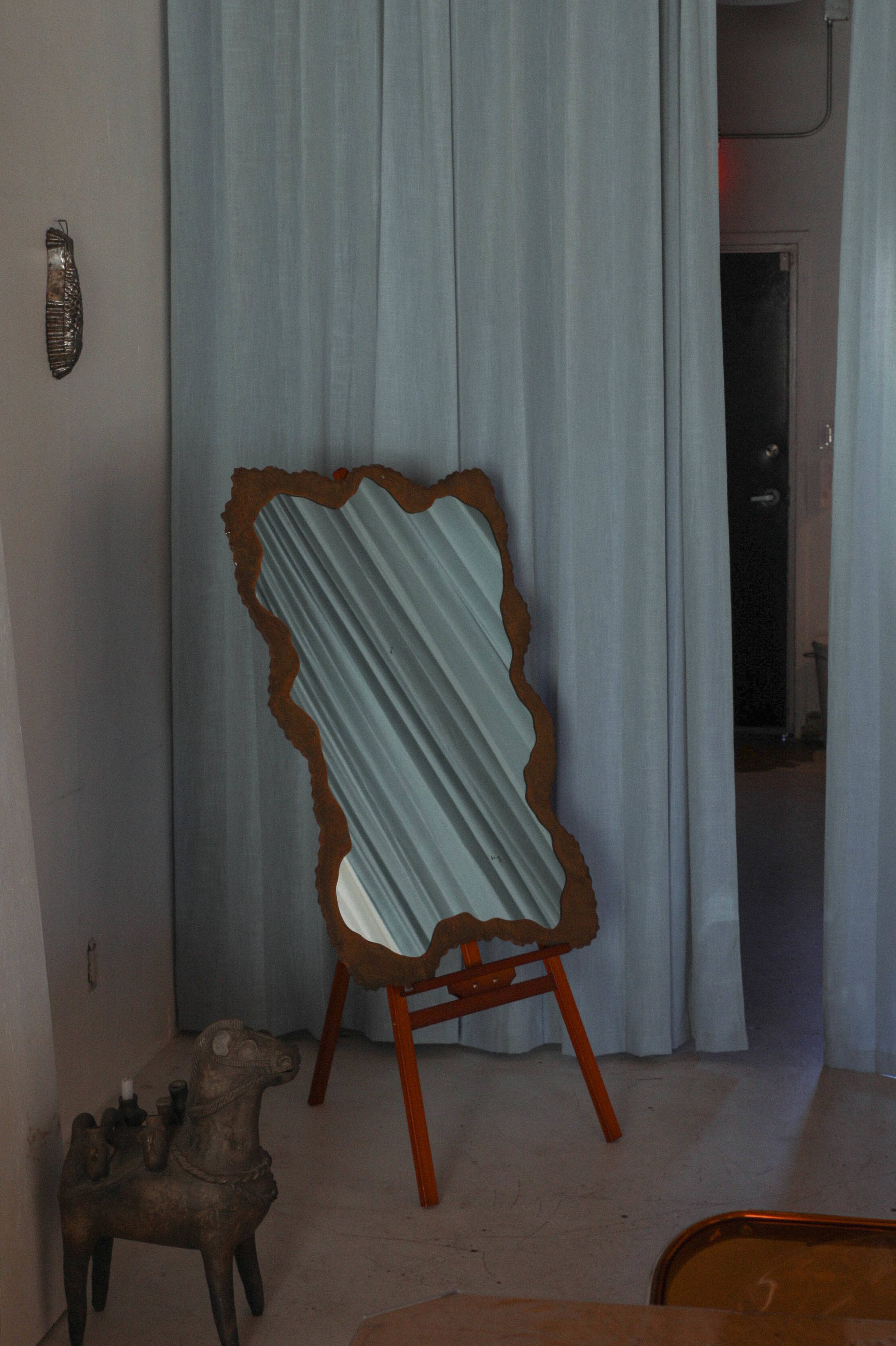 A decorative, wavy-edged mirror stands on a wooden frame near a light blue curtain in a minimal interior space.
