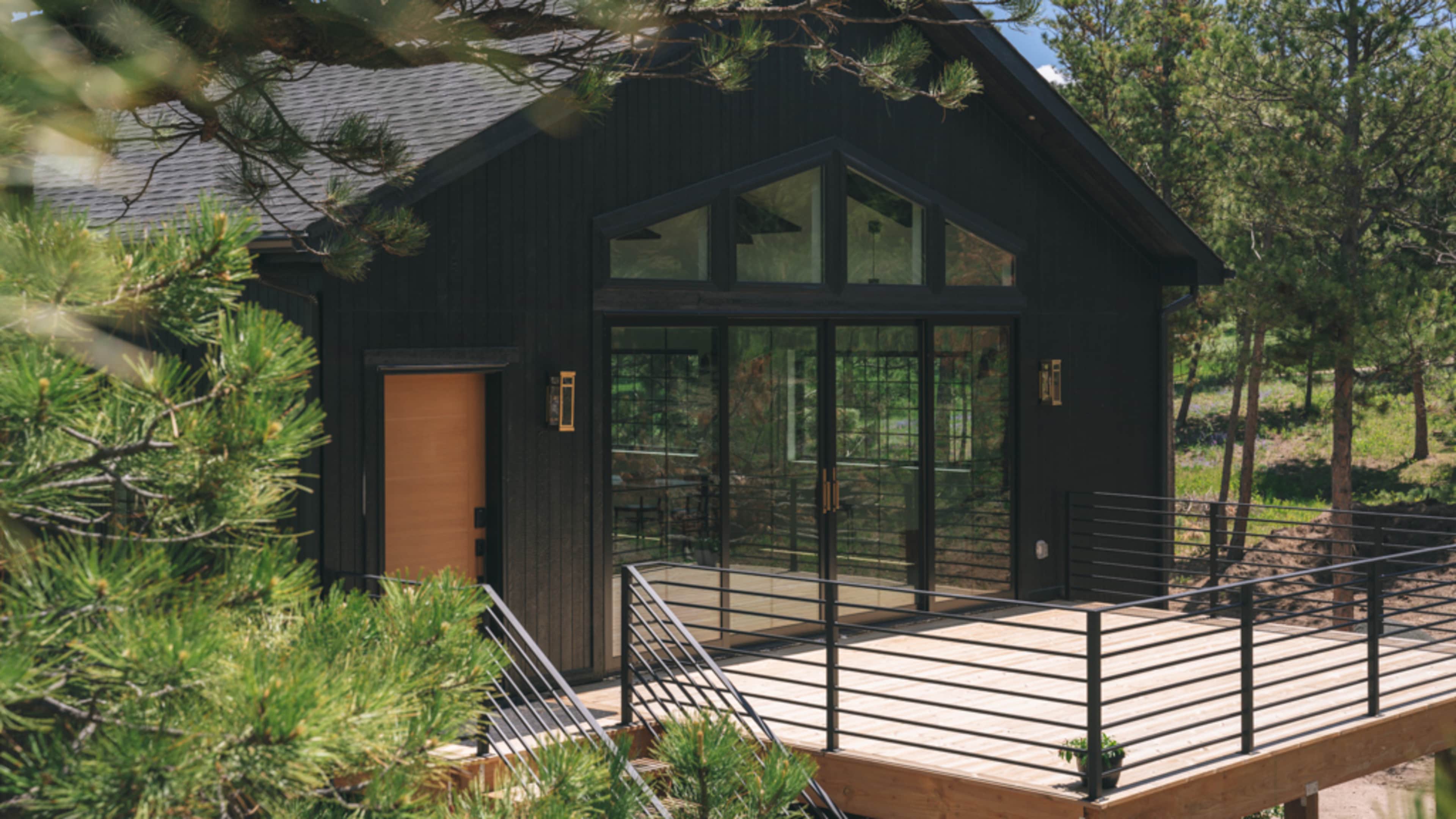 A modern black cabin features large glass doors and a wooden deck, surrounded by greenery.