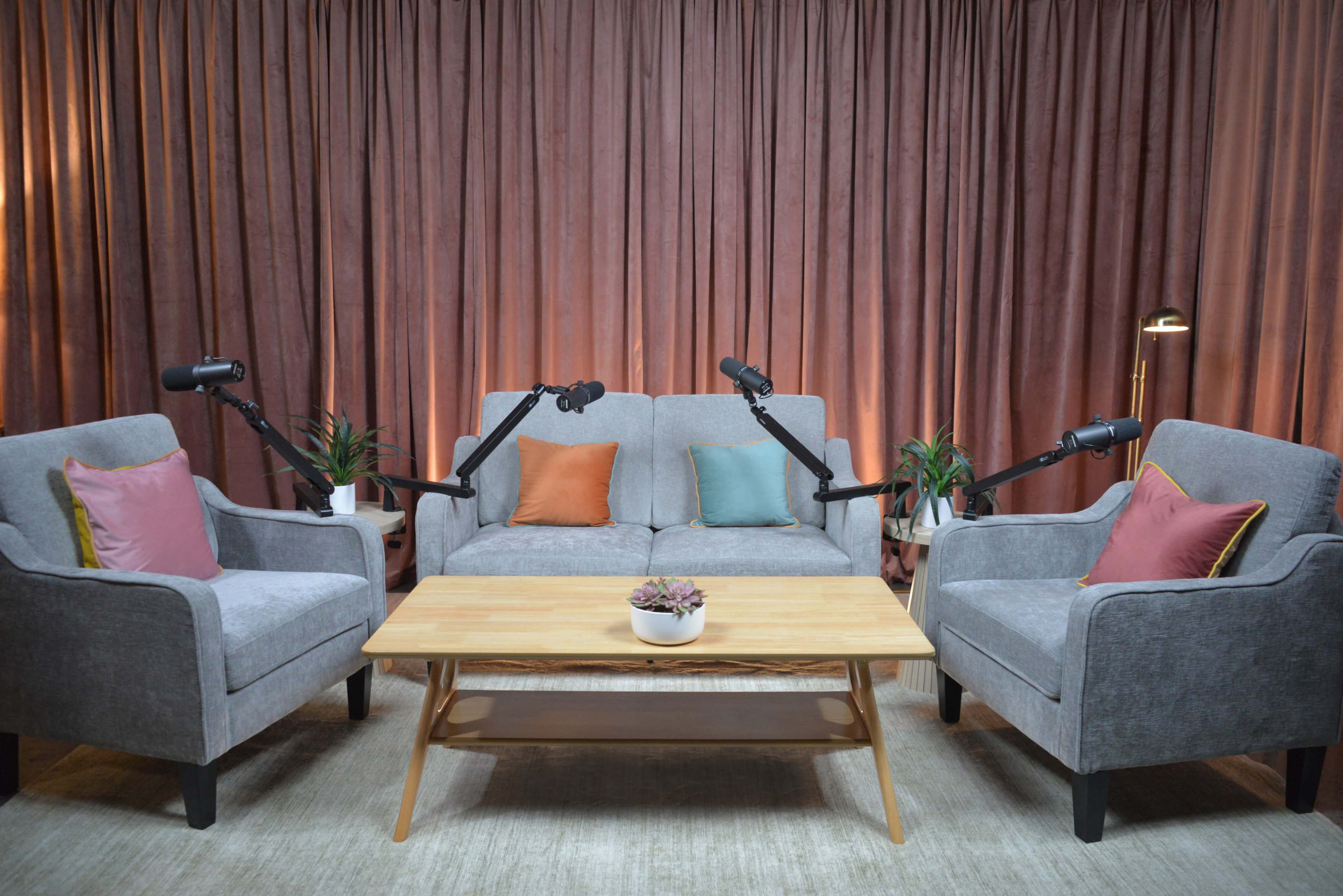 A cozy interview setup features a gray couch with colorful cushions, two armchairs, and a wooden coffee table in front of a backdrop of pink curtains and indoor plants.