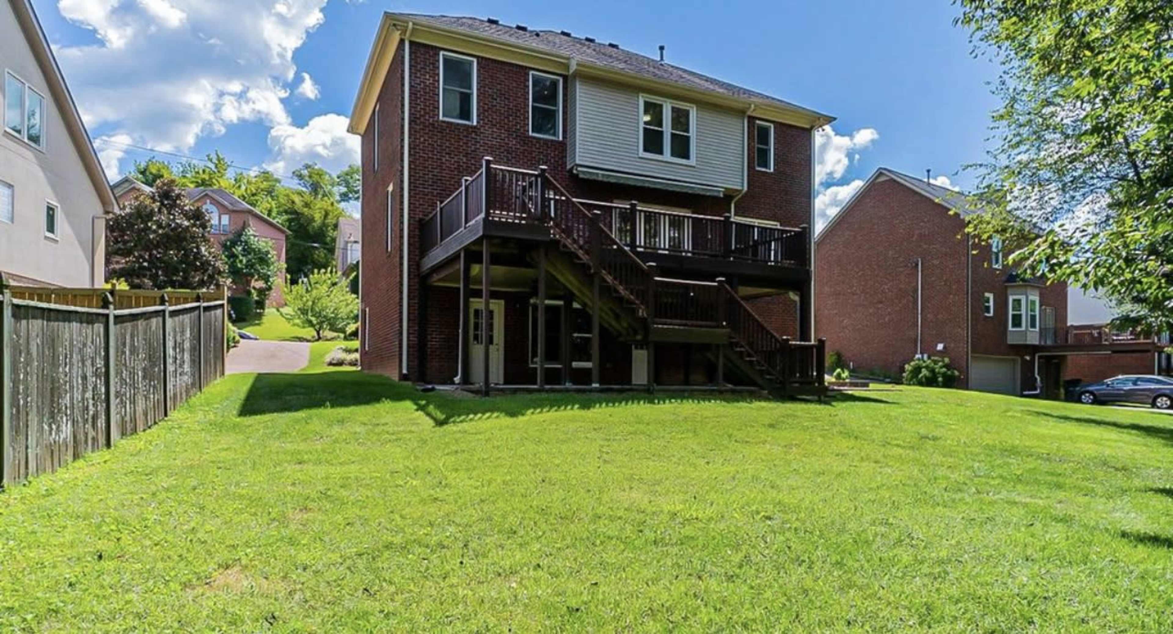 A two-story brick house with a wooden deck and stairs leading to a grassy backyard, surrounded by other residential homes.