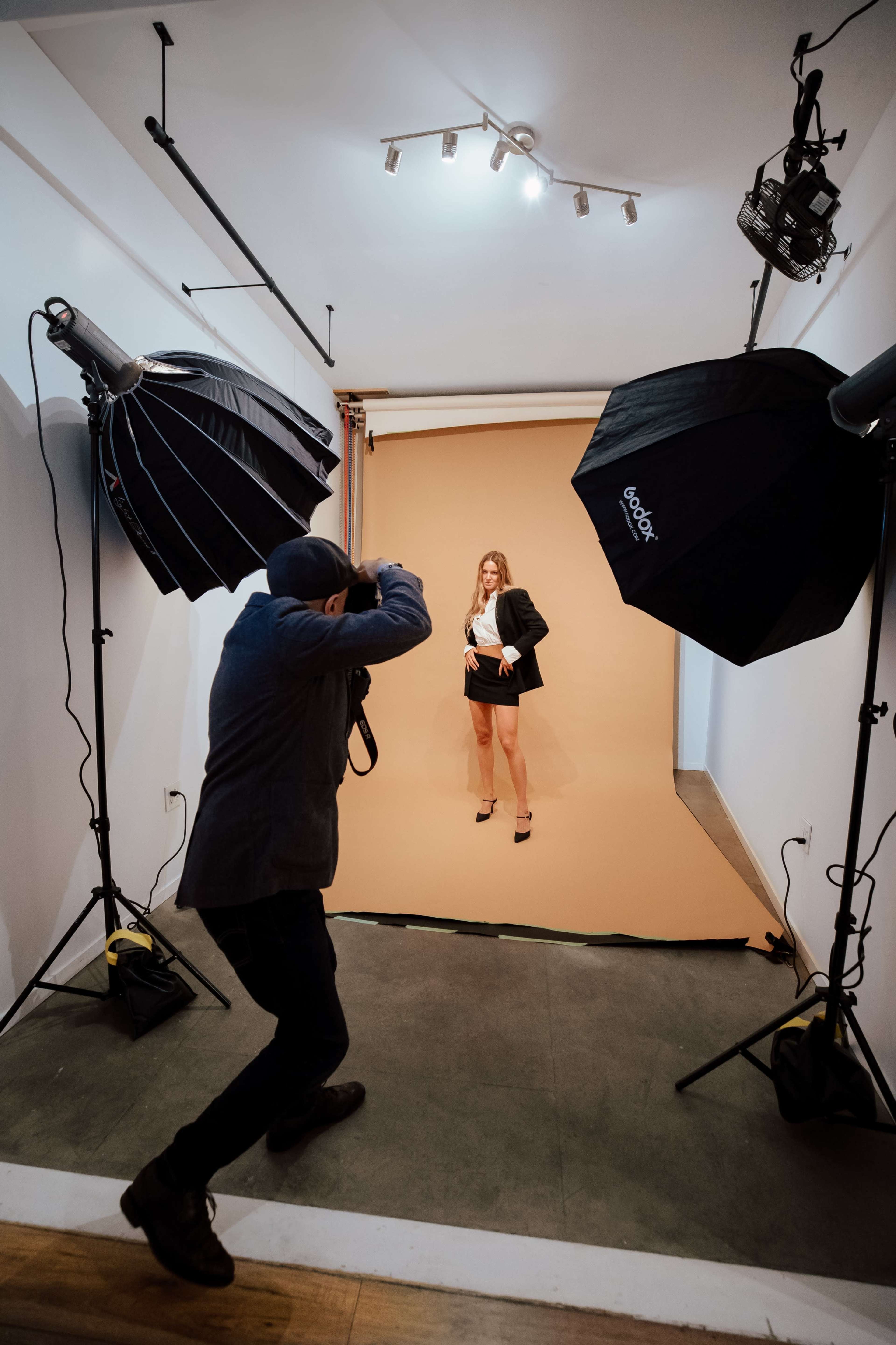 A photographer captures a model posed against a plain beige backdrop in a studio equipped with large lighting equipment.