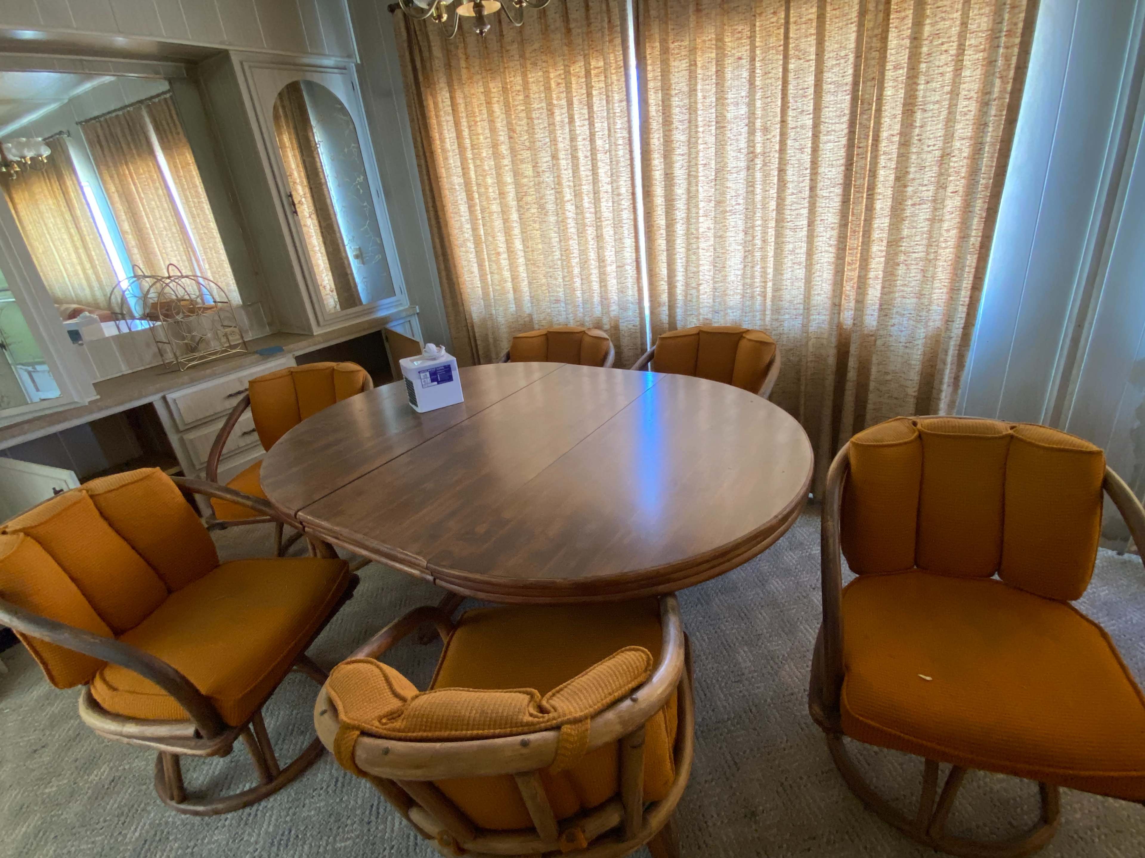 A round wooden dining table is surrounded by six cushioned chairs in a brightly lit room with sheer curtains.