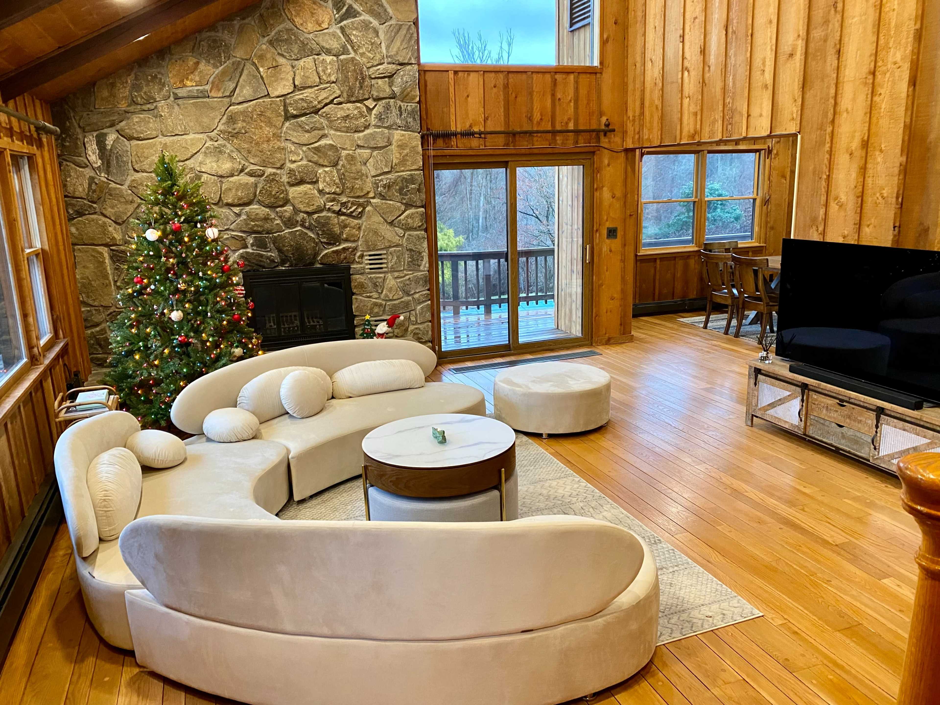 The interior of a wooden cabin features a stone fireplace, a Christmas tree, a circular sofa, and a large television near a dining area with a view of the outdoors.