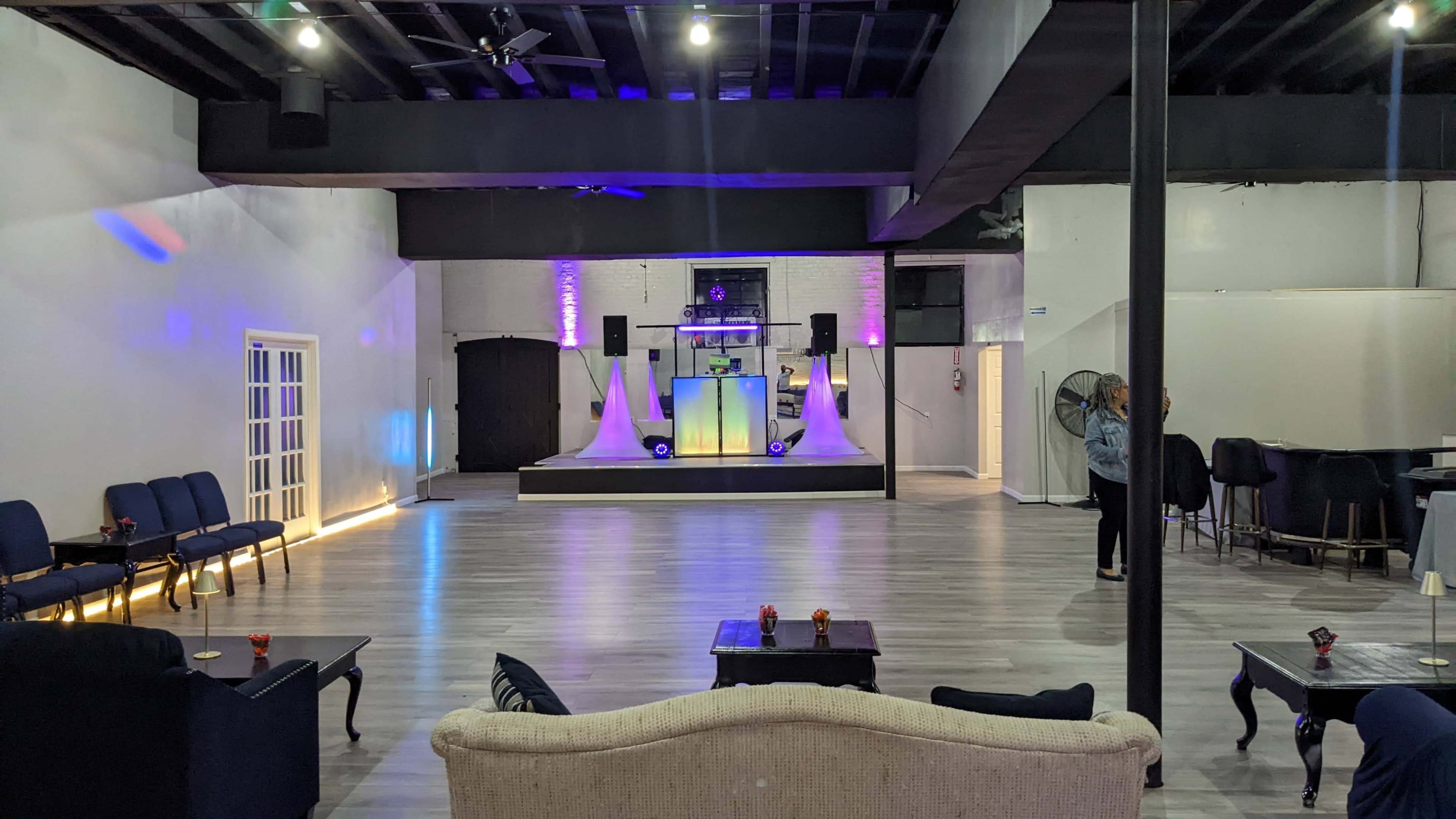A spacious event venue with a stage featuring speakers and decorative lighting, surrounded by seating areas and a bar in the background.