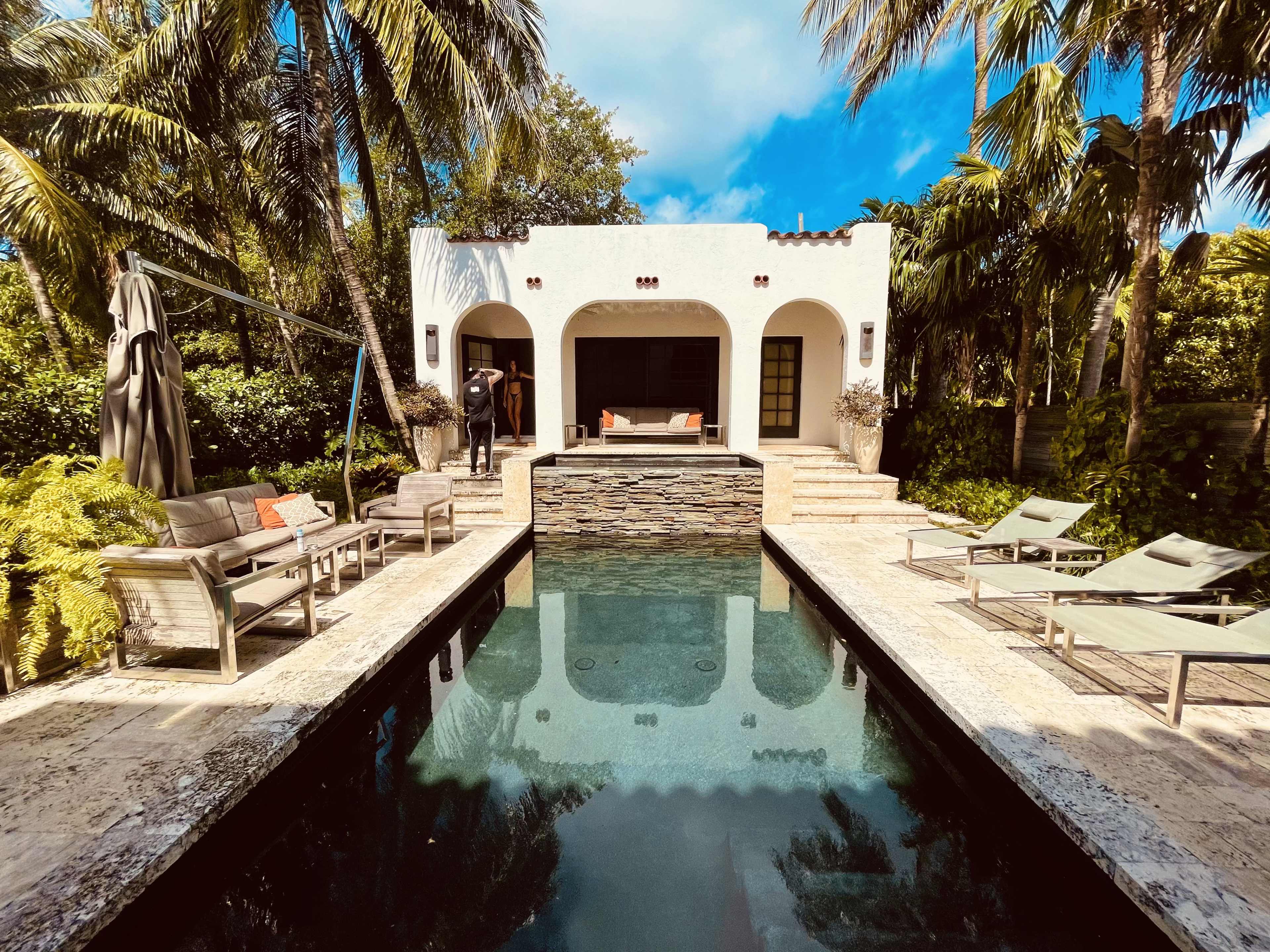 A serene outdoor scene features a rectangular swimming pool surrounded by palm trees, lounge chairs, and a small modern pavilion with a stone accent wall.