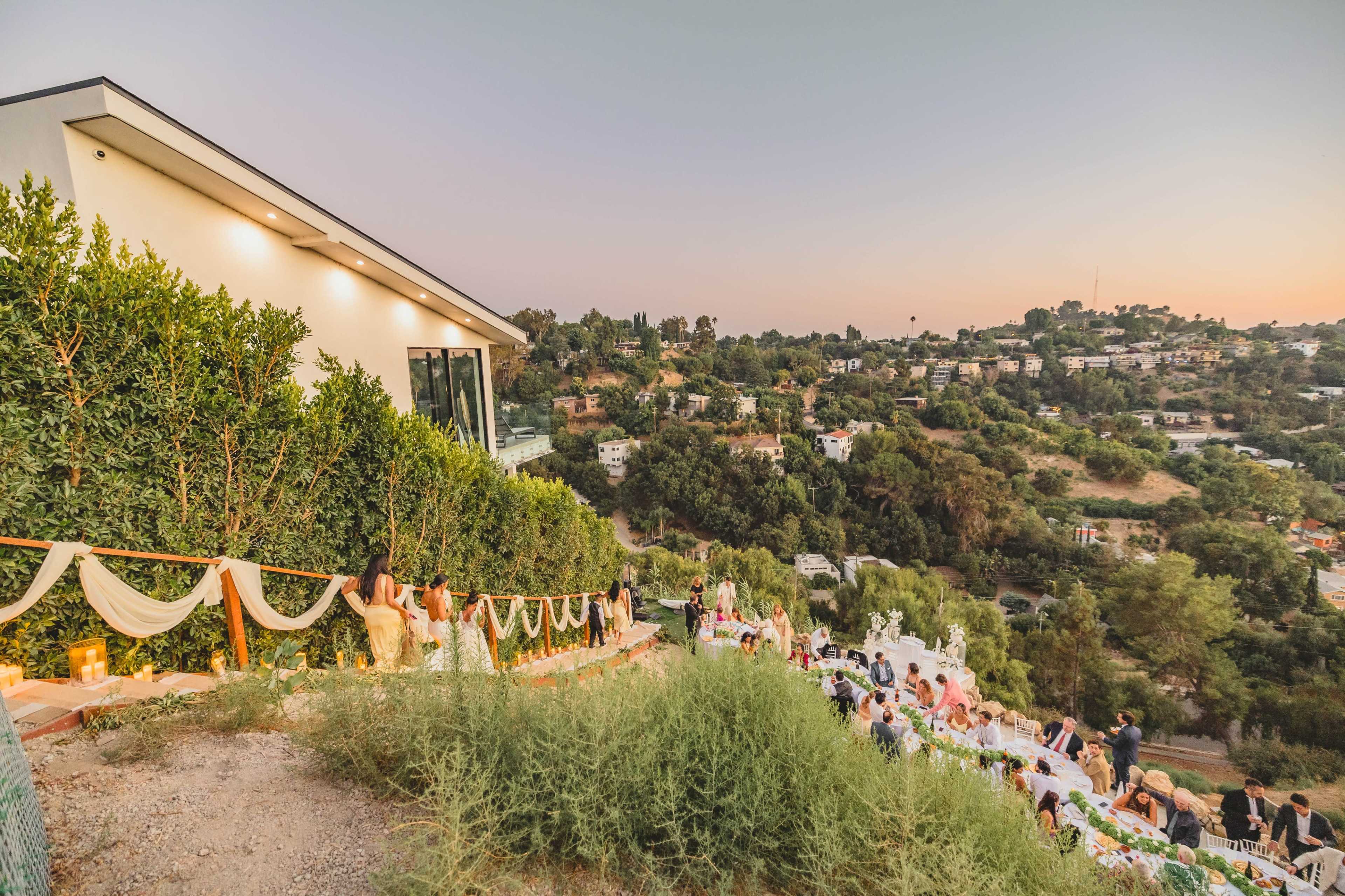 A wedding reception is taking place outdoors on a hillside, featuring tables set up with guests mingling and a scenic view of the surrounding neighborhood at dusk.