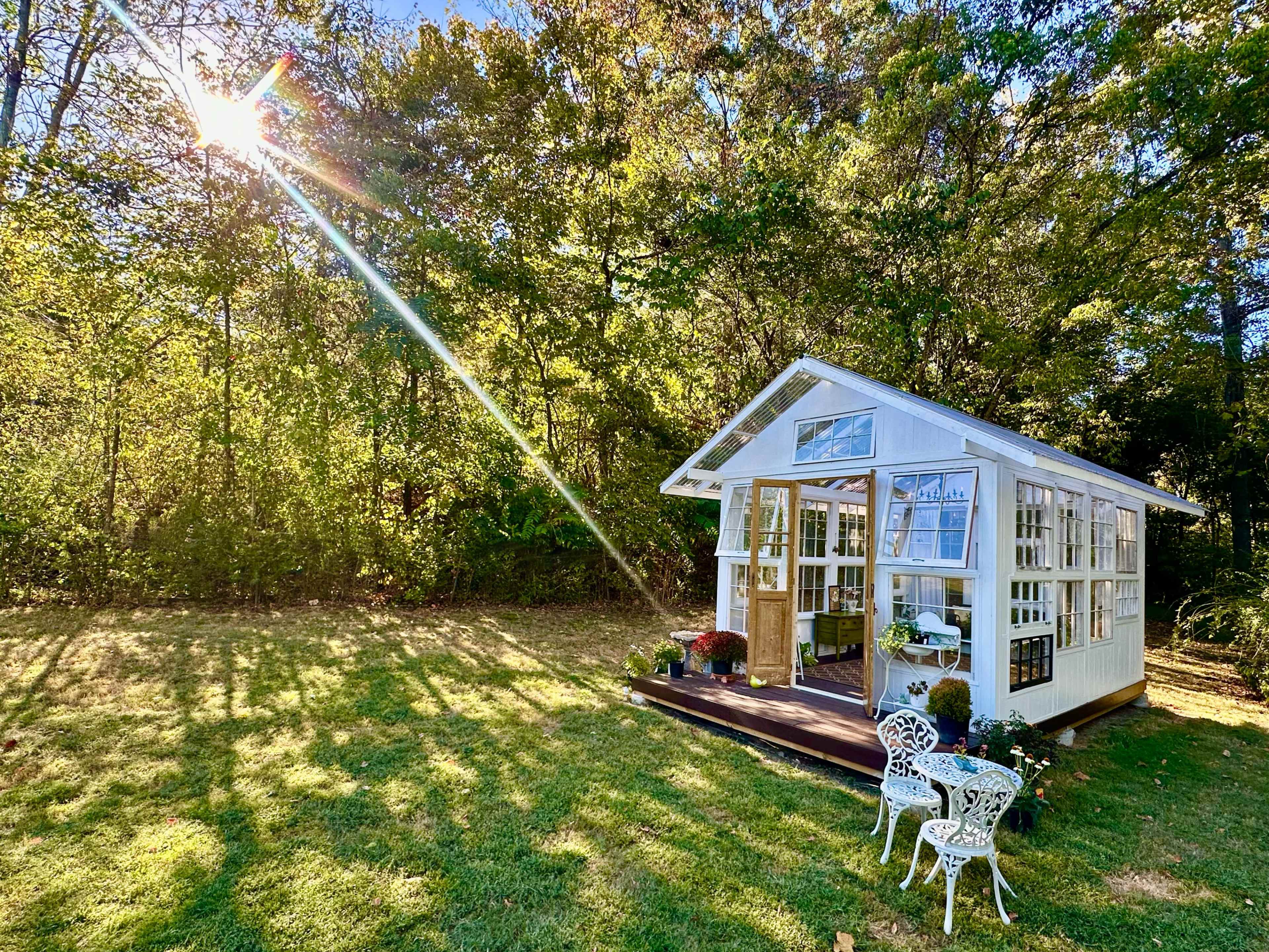 A small white building with large windows stands in a grassy area, surrounded by trees, with sunlight streaming through the branches.