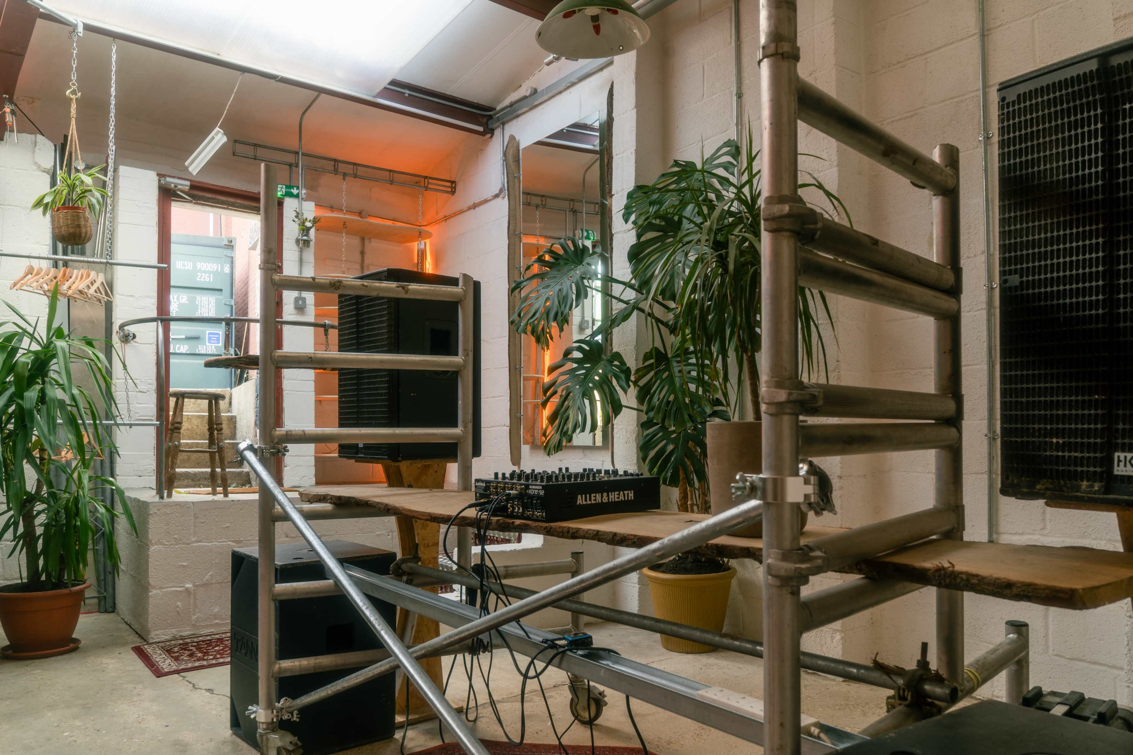 The image shows a room with a scaffolding setup, a mixing console, and several potted plants, illuminated by warm overhead lighting.