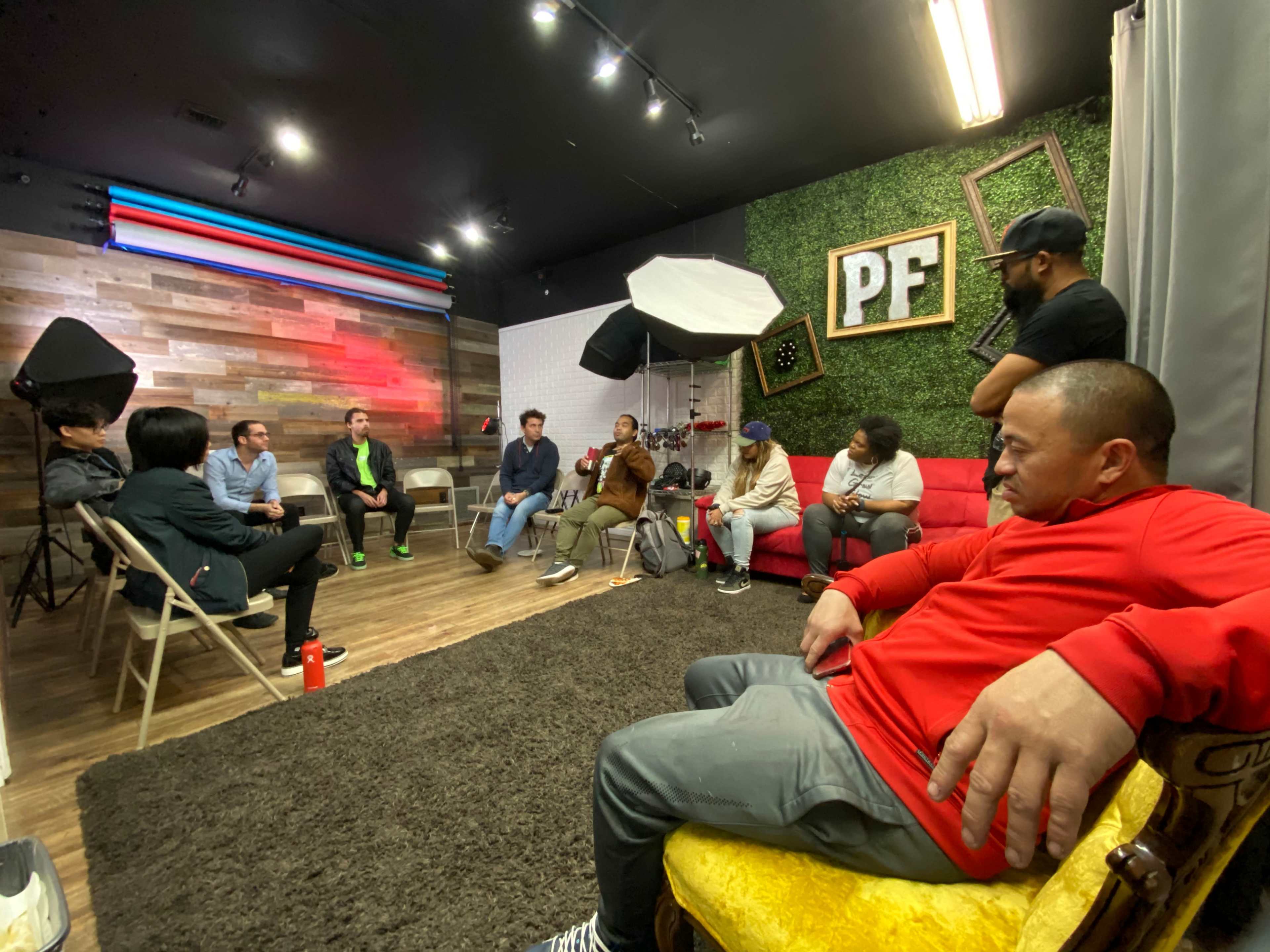 A group of people sits in a circle within a room featuring decorative walls and studio lighting.