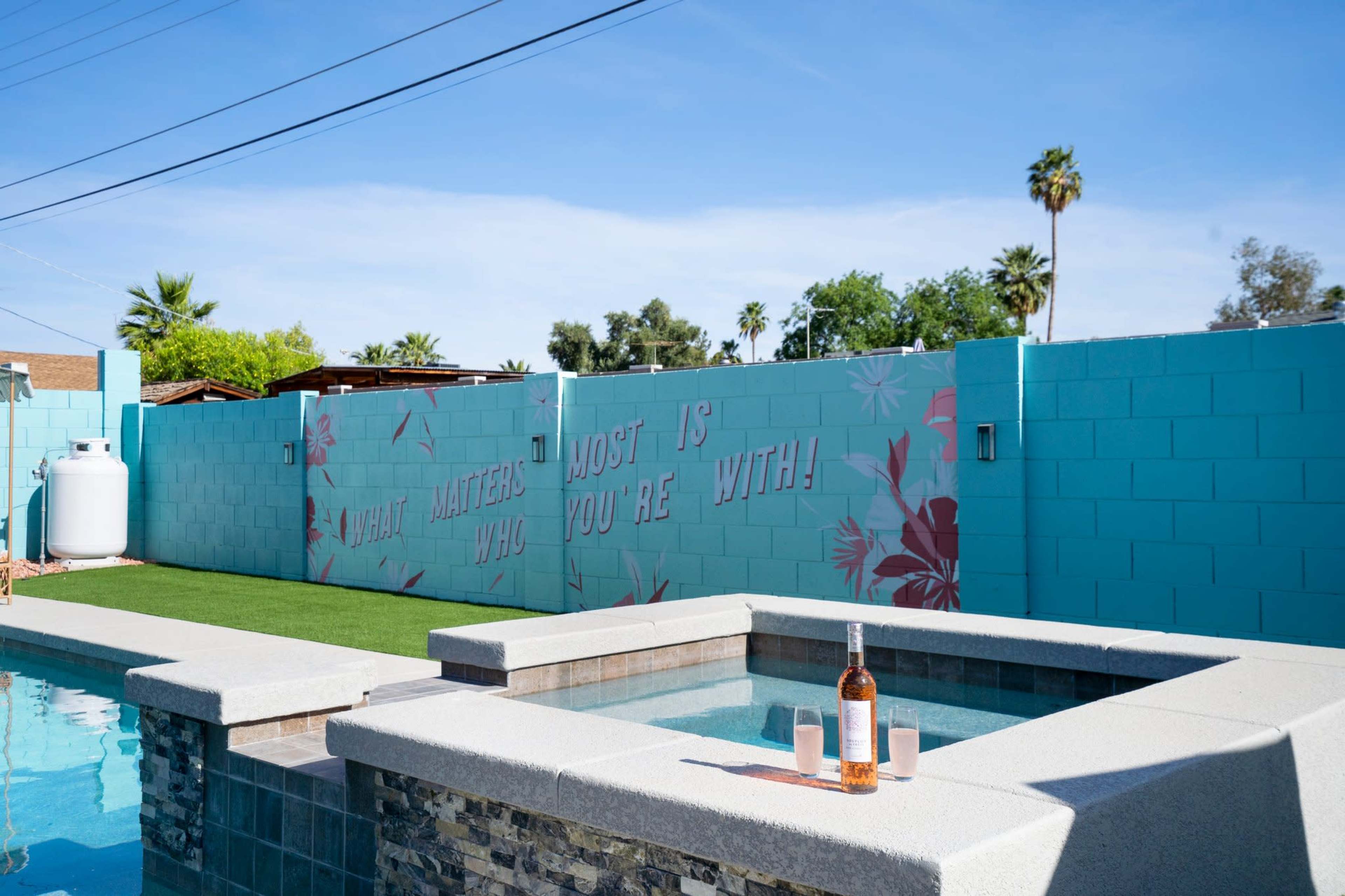 A hot tub sits beside a swimming pool, with a wall behind it featuring the phrase "WHAT MATTERS MOST IS WHO YOU'RE WITH" in stylized lettering, surrounded by palm trees and blue skies.