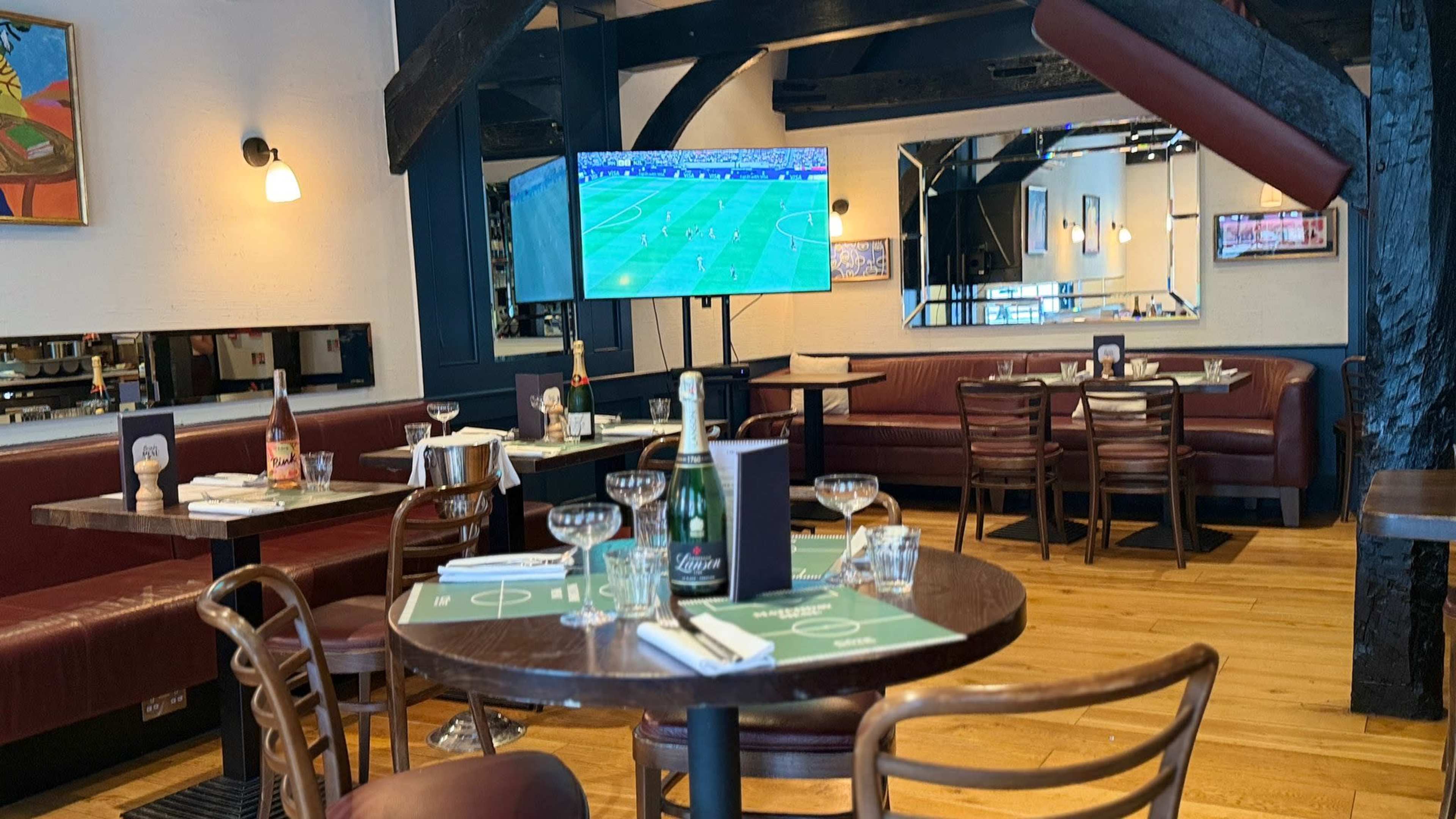 The image shows a restaurant interior featuring wooden beams, a television displaying a sports event, and tables with chairs arranged around them.
