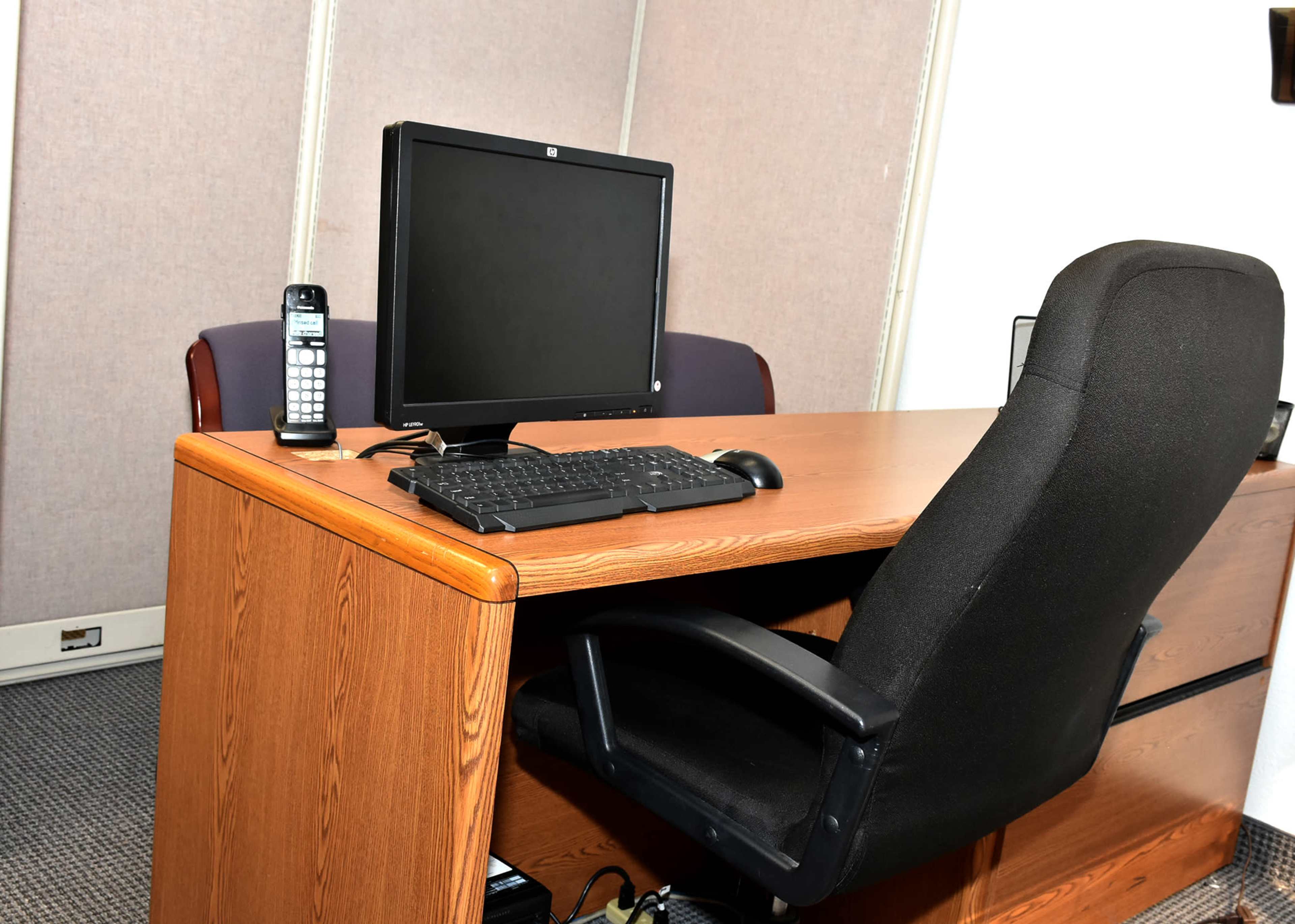 An office setup with a wooden desk, an empty black office chair, a desktop computer monitor, a keyboard, and a telephone.