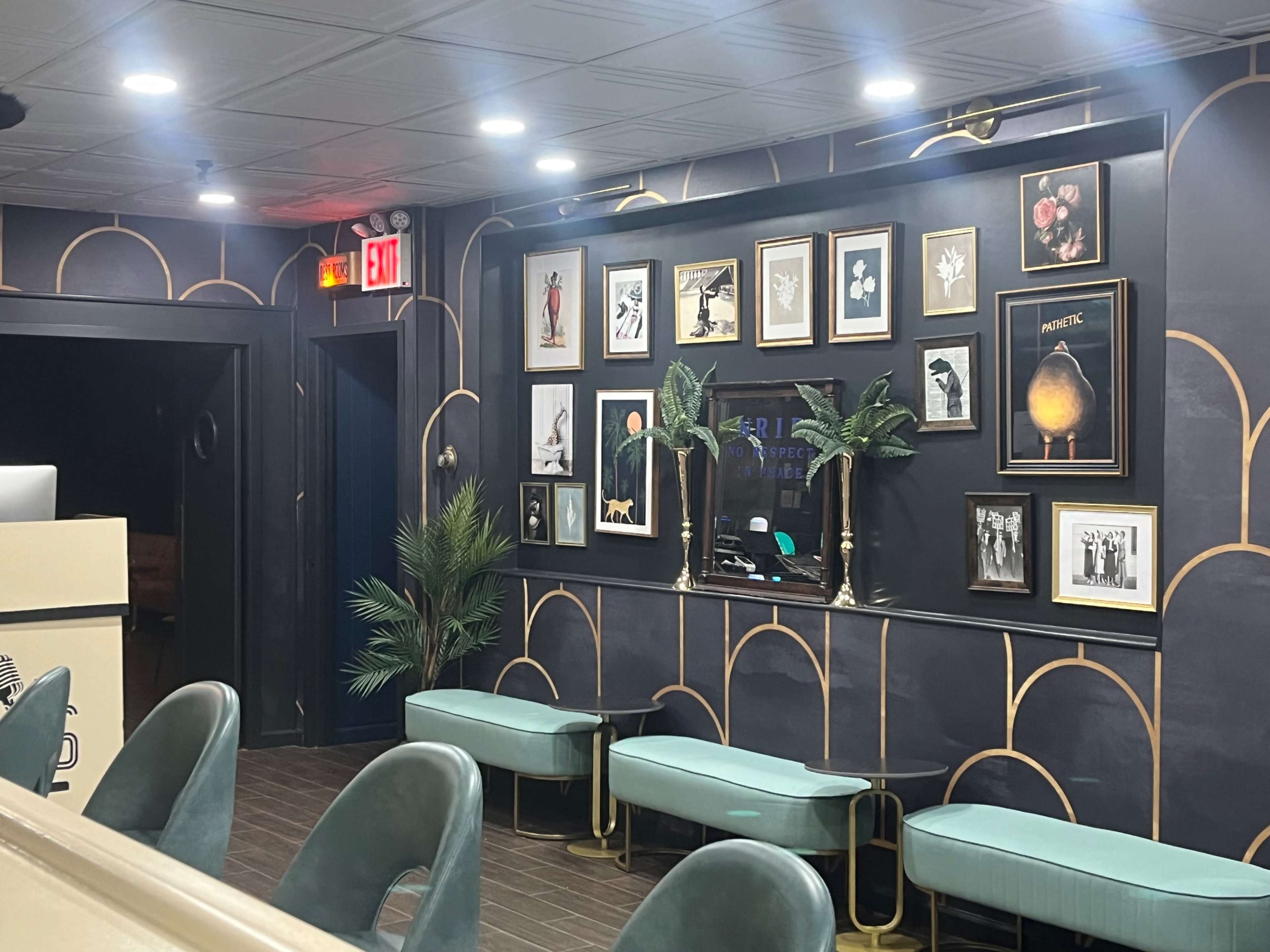 A modern waiting area featuring green upholstered seating, decorative wall frames with artwork, and a potted plant against a dark patterned wall.