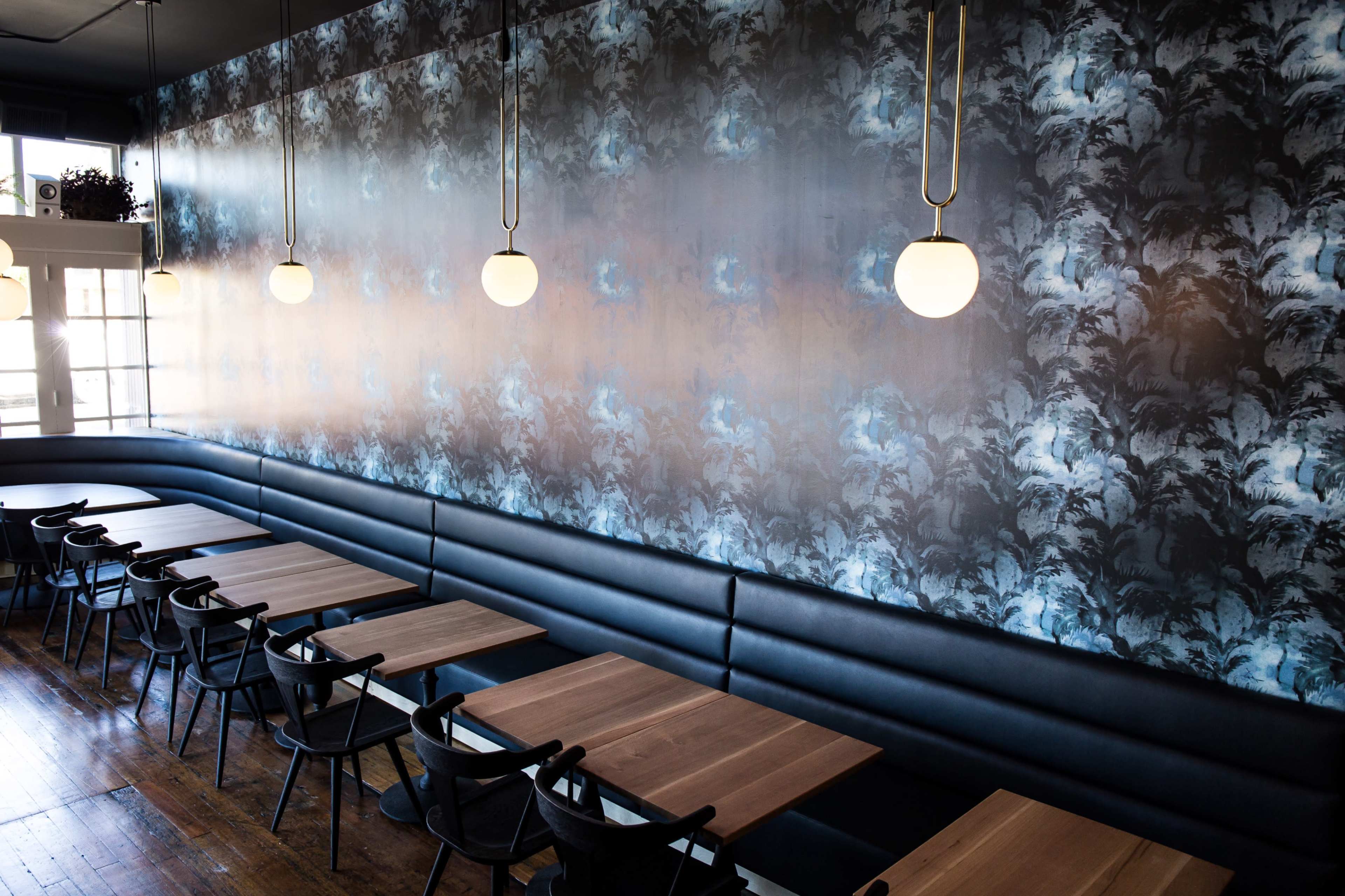 A dining area with a patterned wallpaper, wooden tables, and black seating along the walls, illuminated by round pendant lights.