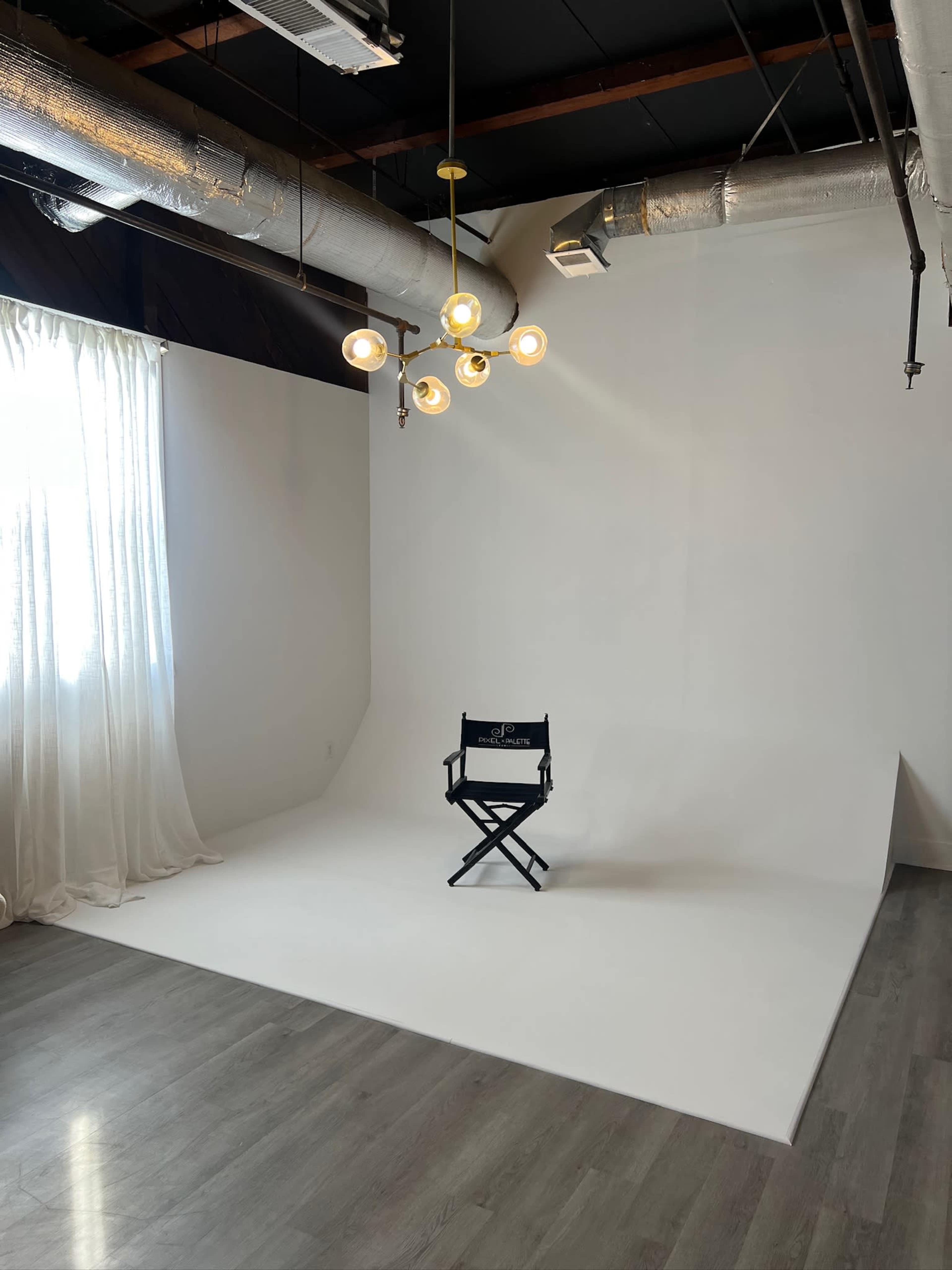 A minimalistic studio space featuring a black director's chair on a smooth white backdrop, with large windows draped in sheer curtains and exposed ductwork overhead.