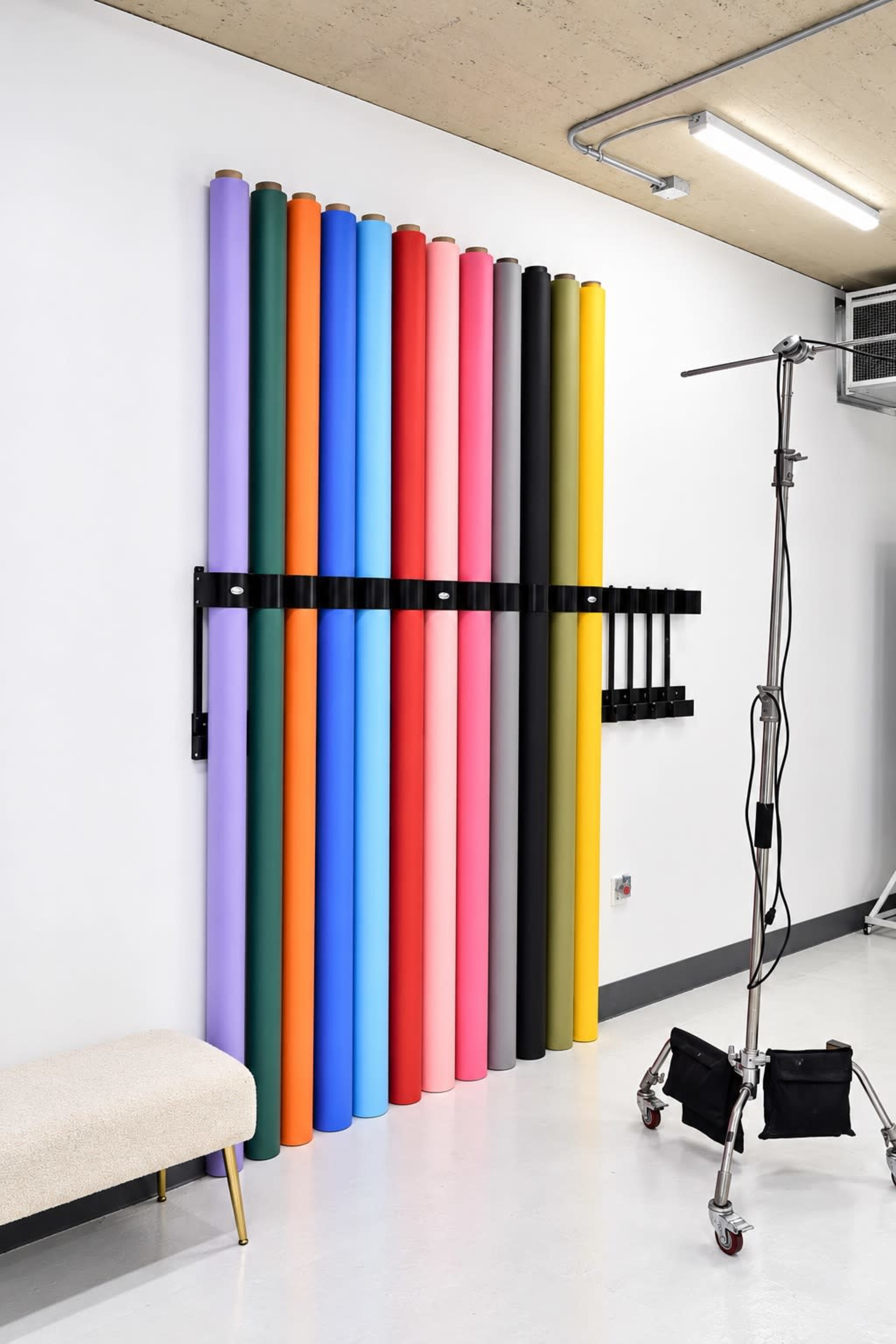 The image shows a wall-mounted display of colorful roll-backdrops alongside a photography lighting stand in a studio environment.