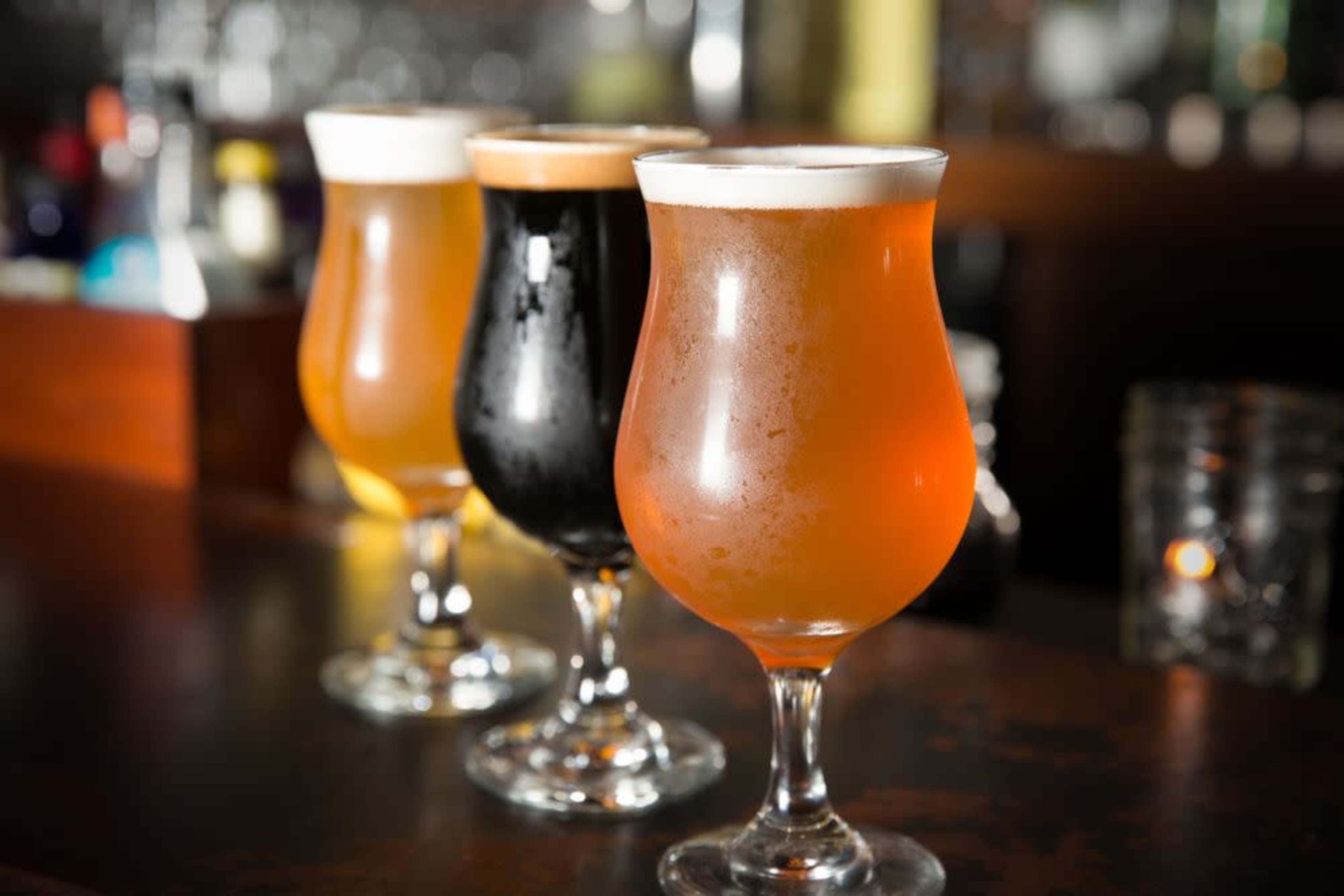 Three glasses of beer filled with different colored brews stand on a bar, showcasing a range of hues from golden to black.