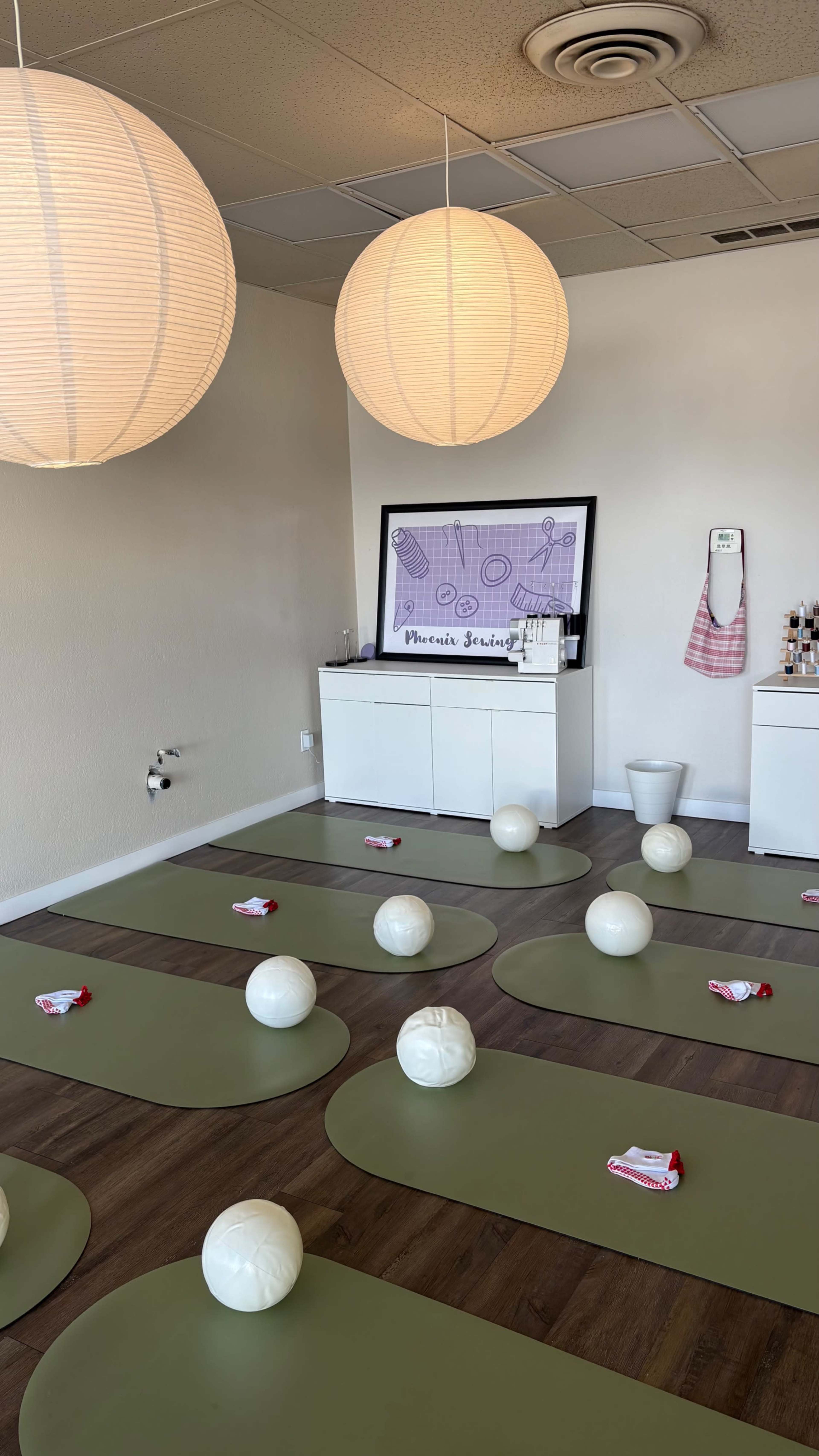 The image shows a yoga studio with green mats arranged on the floor, white exercise balls scattered around, and soft pendant lights hanging from the ceiling.