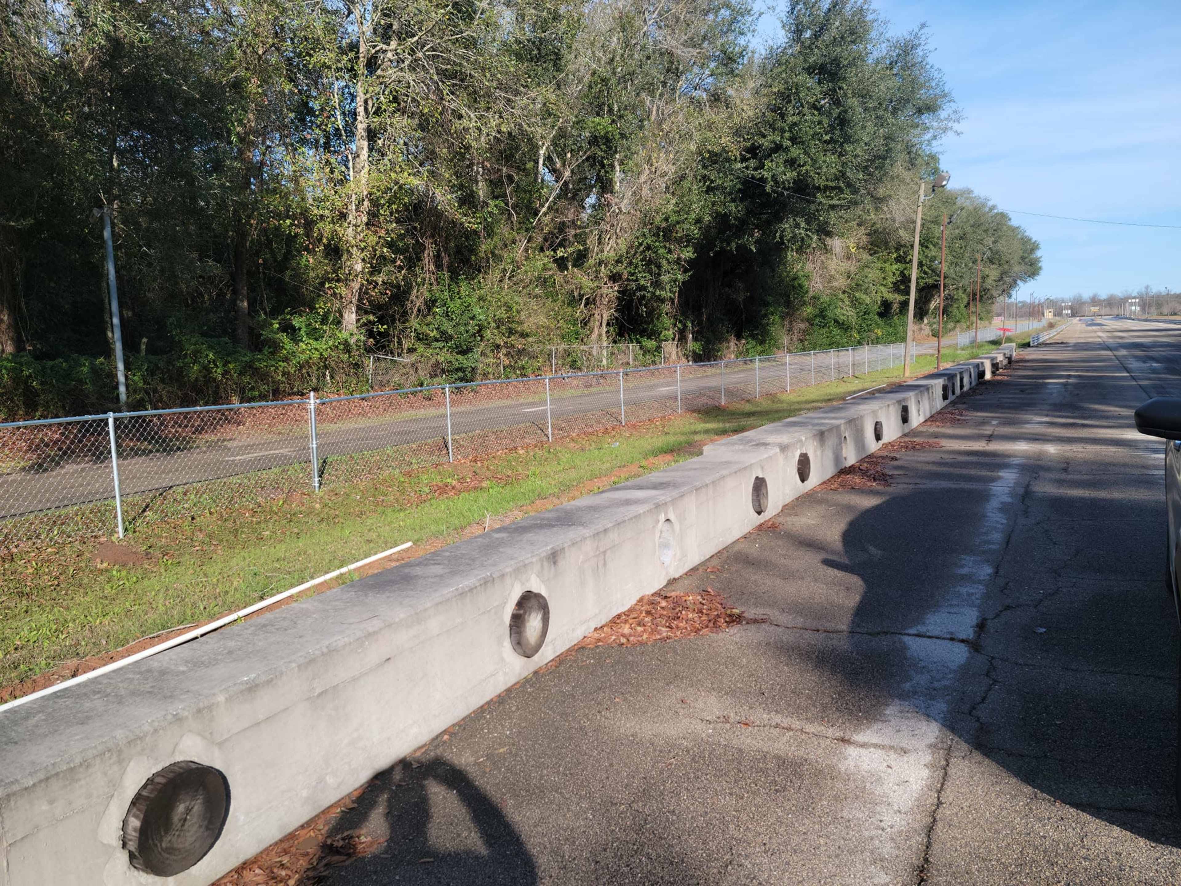 The image shows a concrete barrier with circular openings beside a paved road and a chain-link fence, surrounded by trees and grass.
