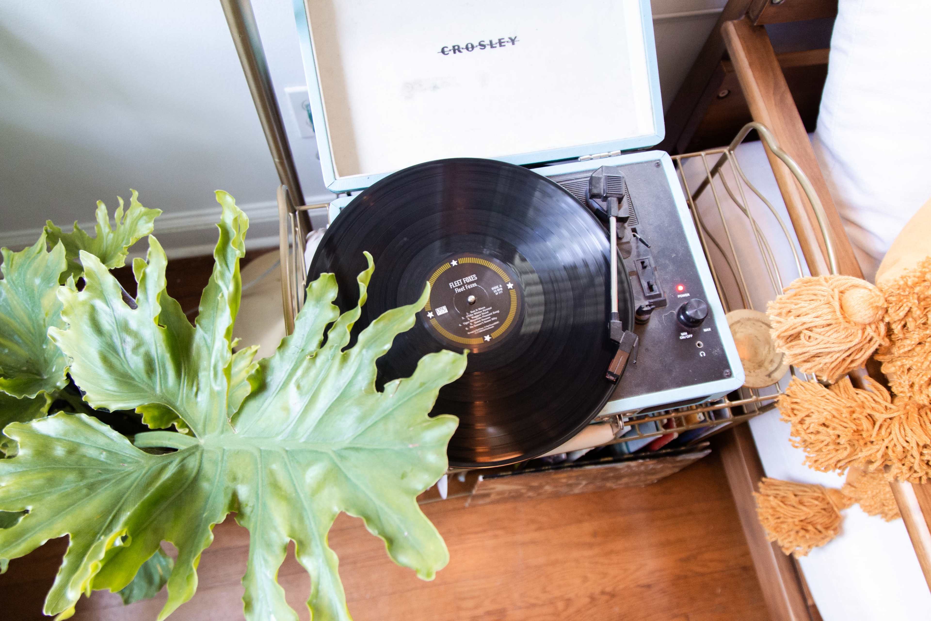 A vinyl record is playing on a Crosley turntable positioned on a wooden shelf next to a large green leaf and a decorative item.