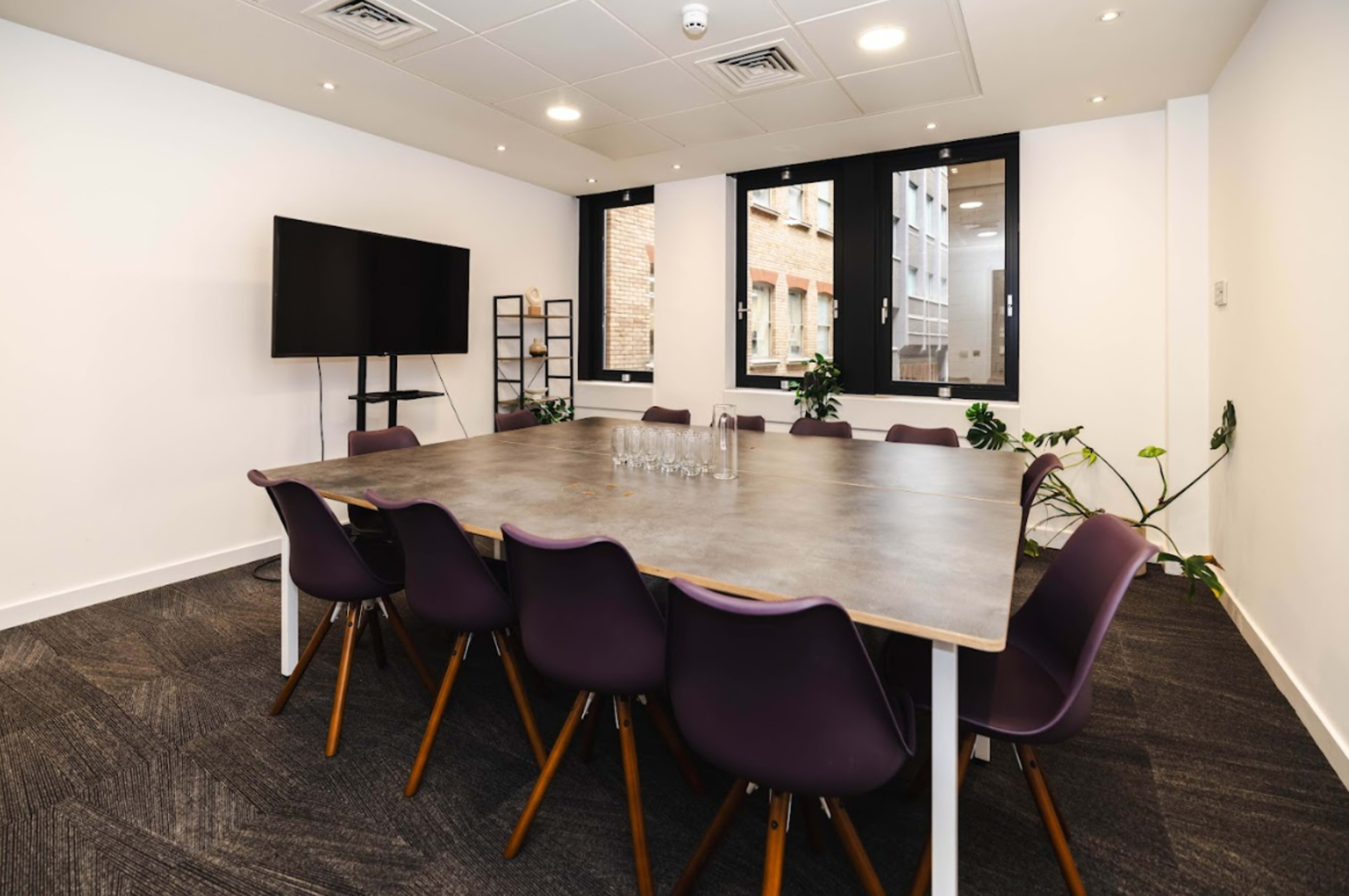 A modern conference room features a large table surrounded by eight purple chairs, with a television mounted on the wall and windows allowing natural light.