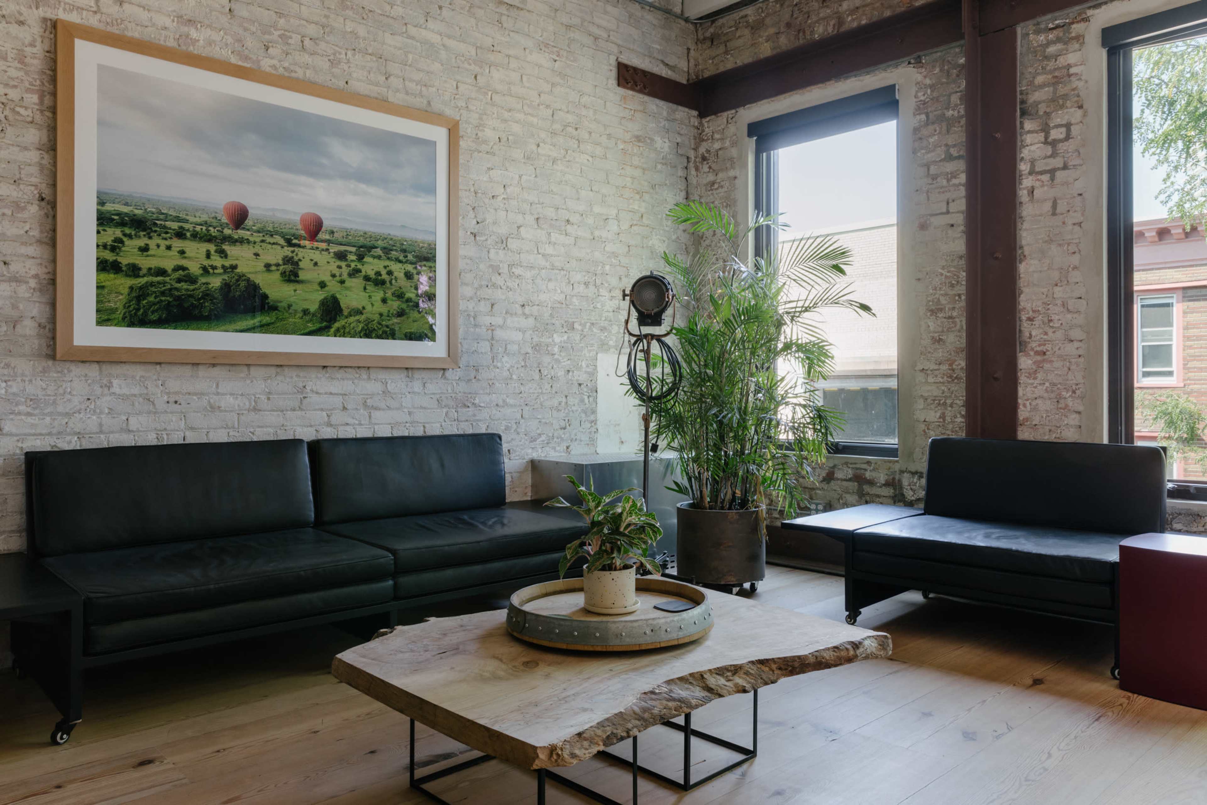 A modern living space with a large framed landscape photograph, a black leather sofa, a wood coffee table, and plants in a well-lit room with exposed brick walls and industrial elements.