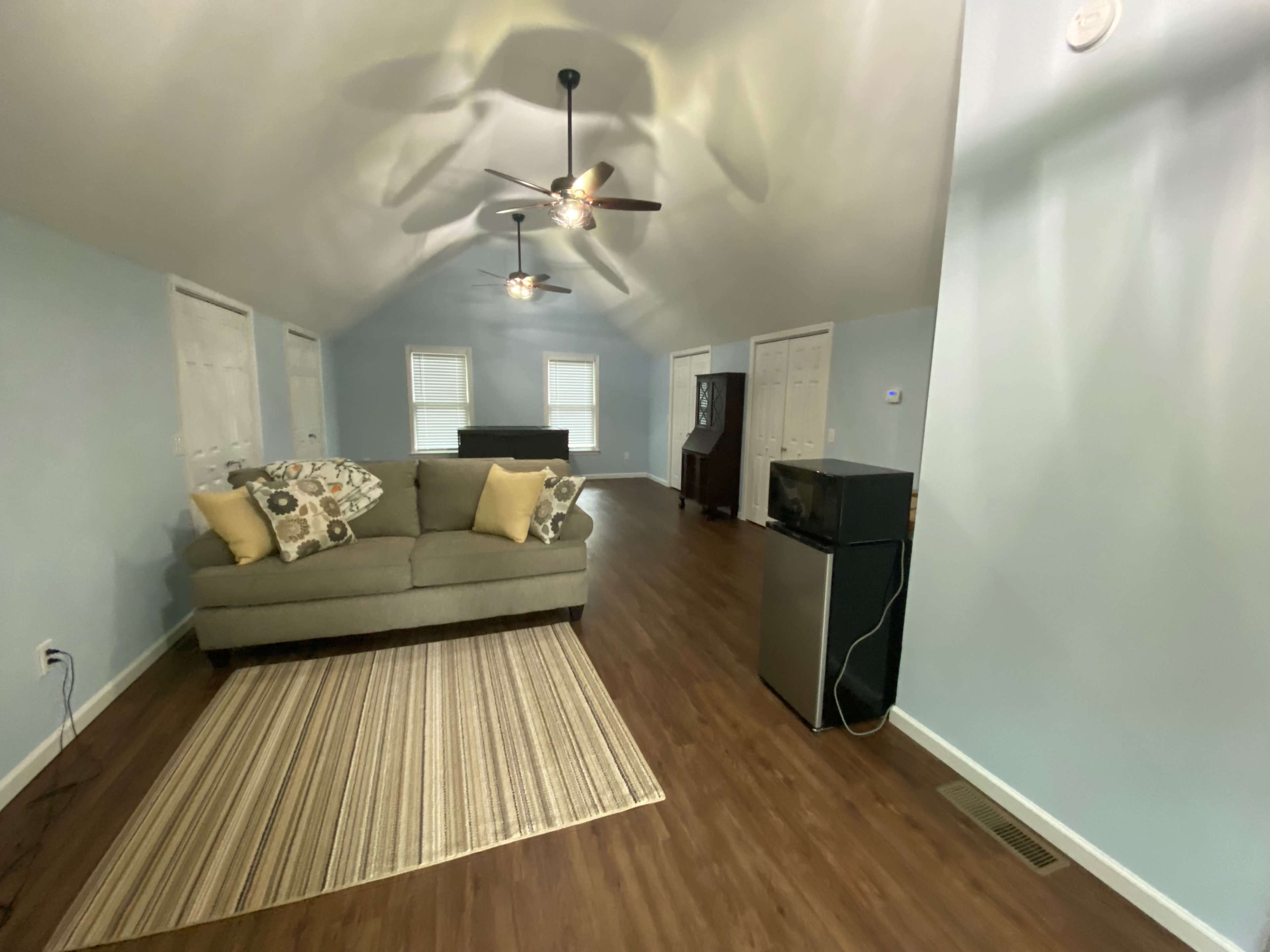 The image shows a light blue living room with a couch, a rug, a small refrigerator, and several doors along the walls.