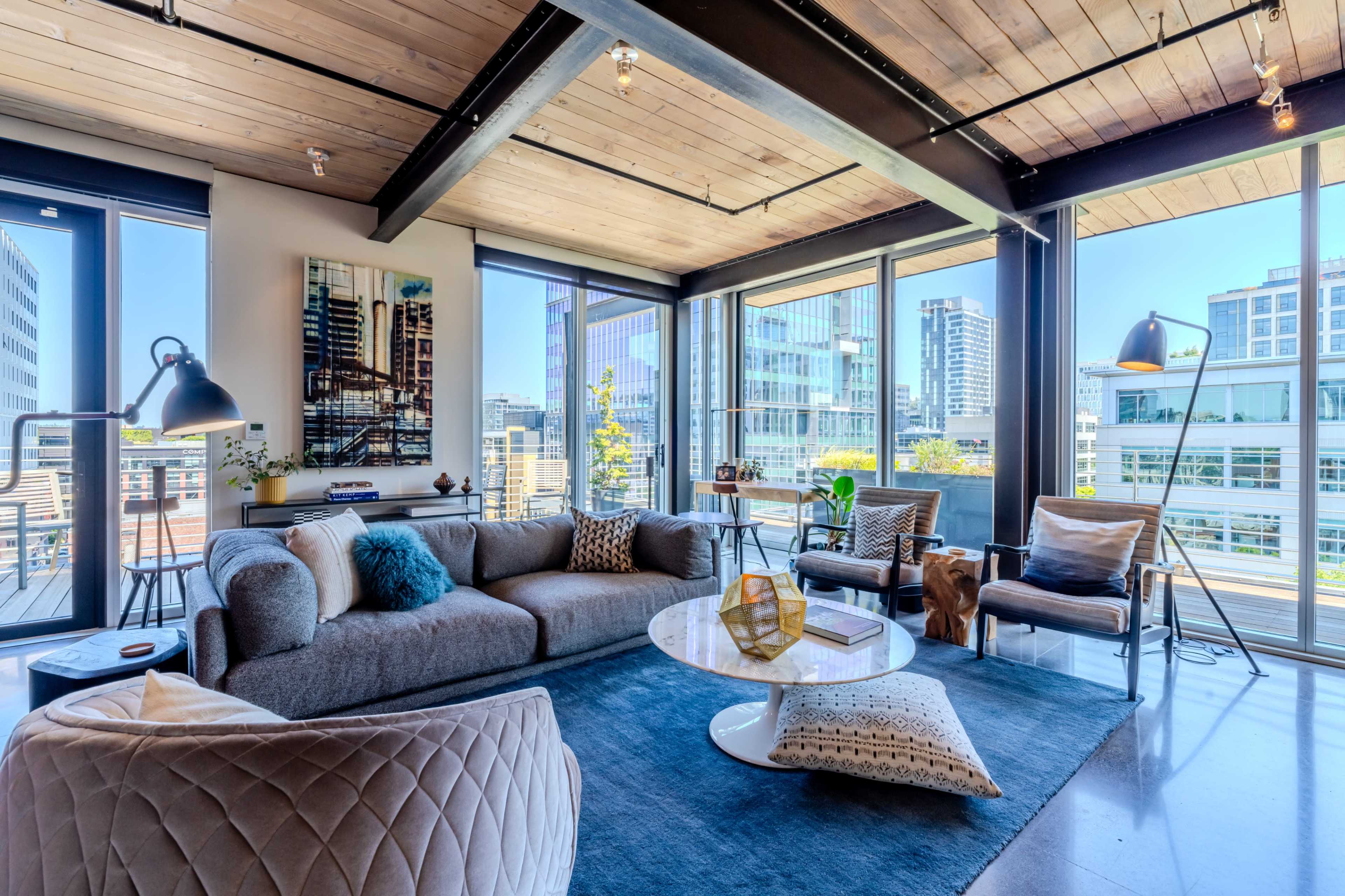 A modern living room features a gray sectional sofa, a round coffee table, and large windows showcasing a city skyline.