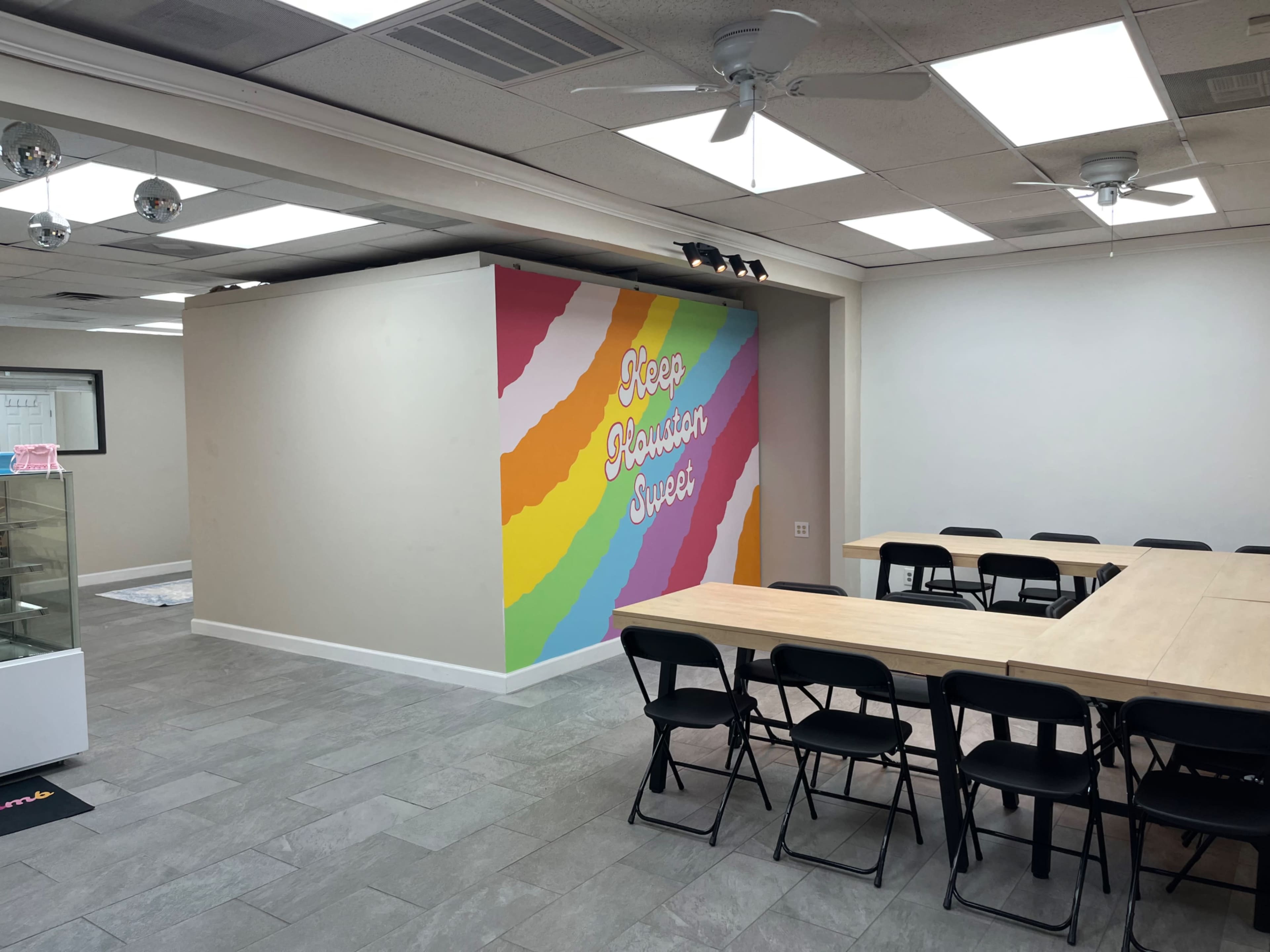The image shows a brightly colored mural with the text "Keep Houston Sweet" on a wall in a spacious room featuring a long wooden table and black chairs.