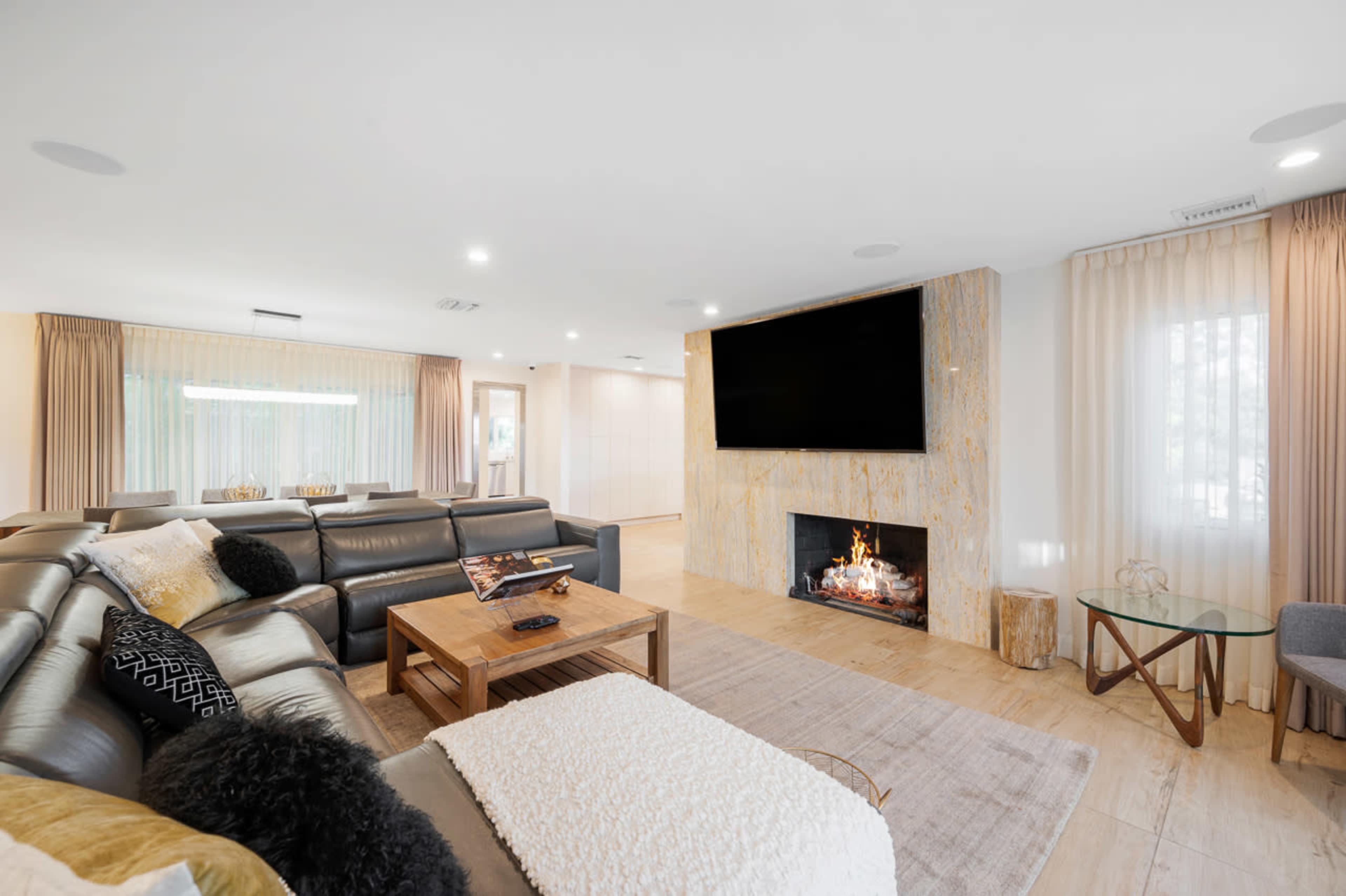 A modern living room features a large sectional sofa, a coffee table, a flat-screen TV mounted above a fireplace, and large windows with sheer curtains.