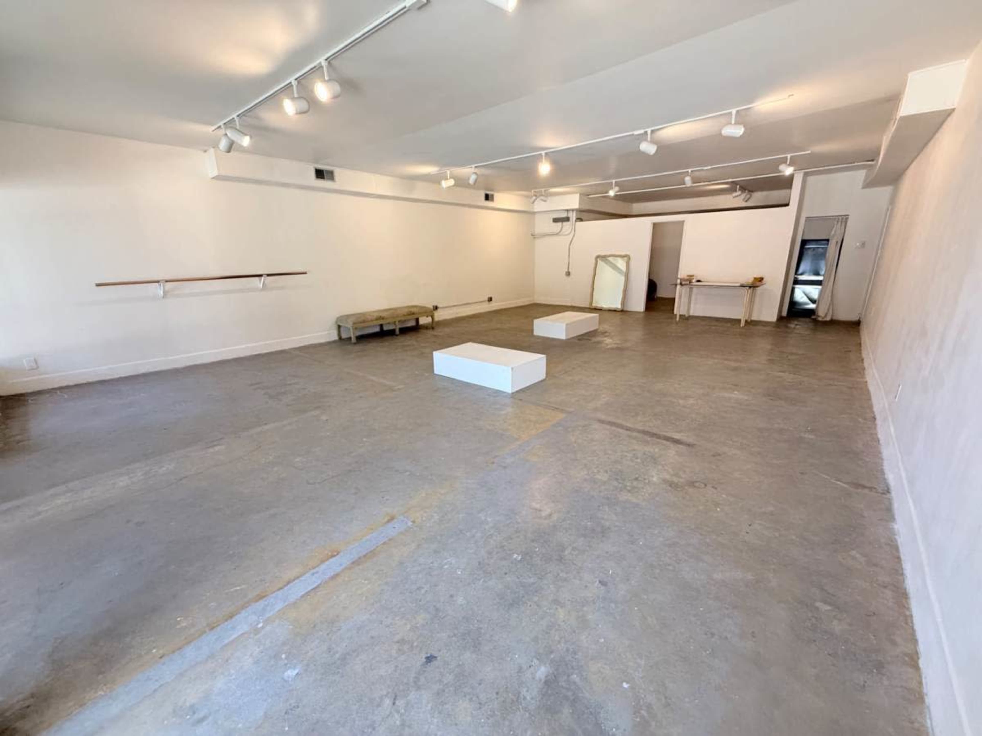 The image shows a spacious, empty room with concrete flooring, white walls, and minimal lighting fixtures.