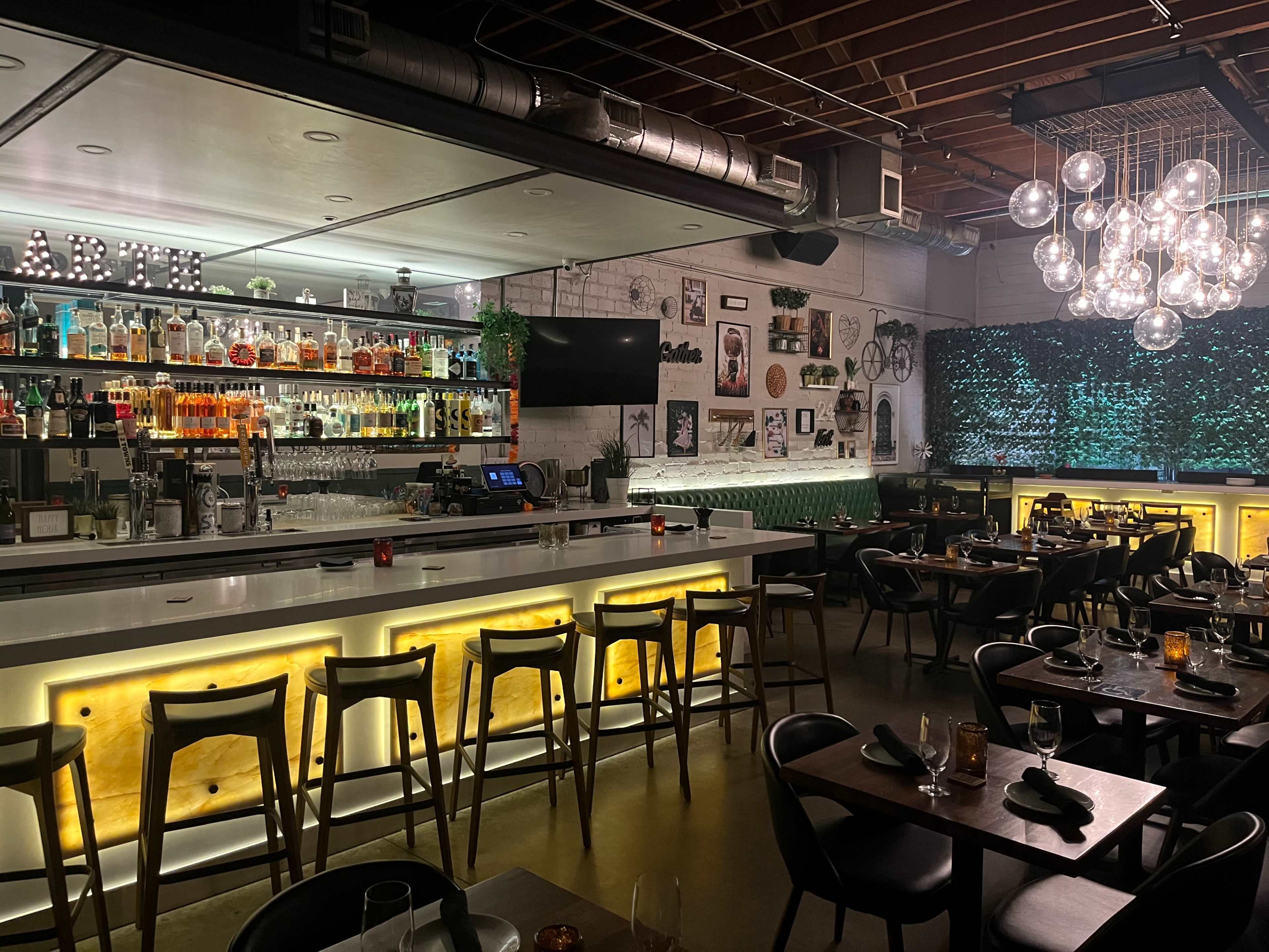 The image shows a modern restaurant interior with a well-lit bar area, stylish seating, and decorative lighting fixtures.