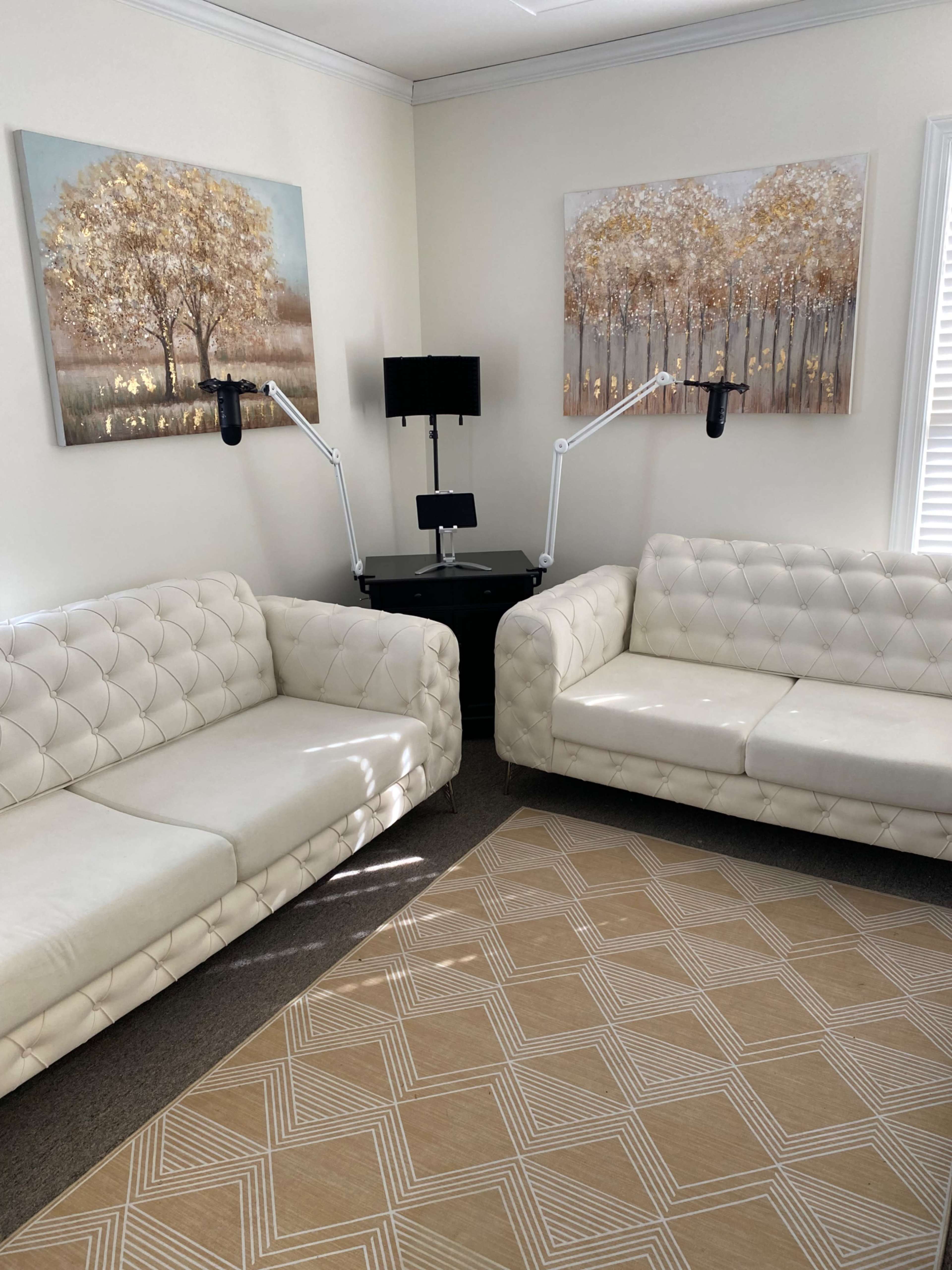 A well-lit room featuring two white tufted couches, a round table with a black stand, and two art pieces depicting trees on the walls.