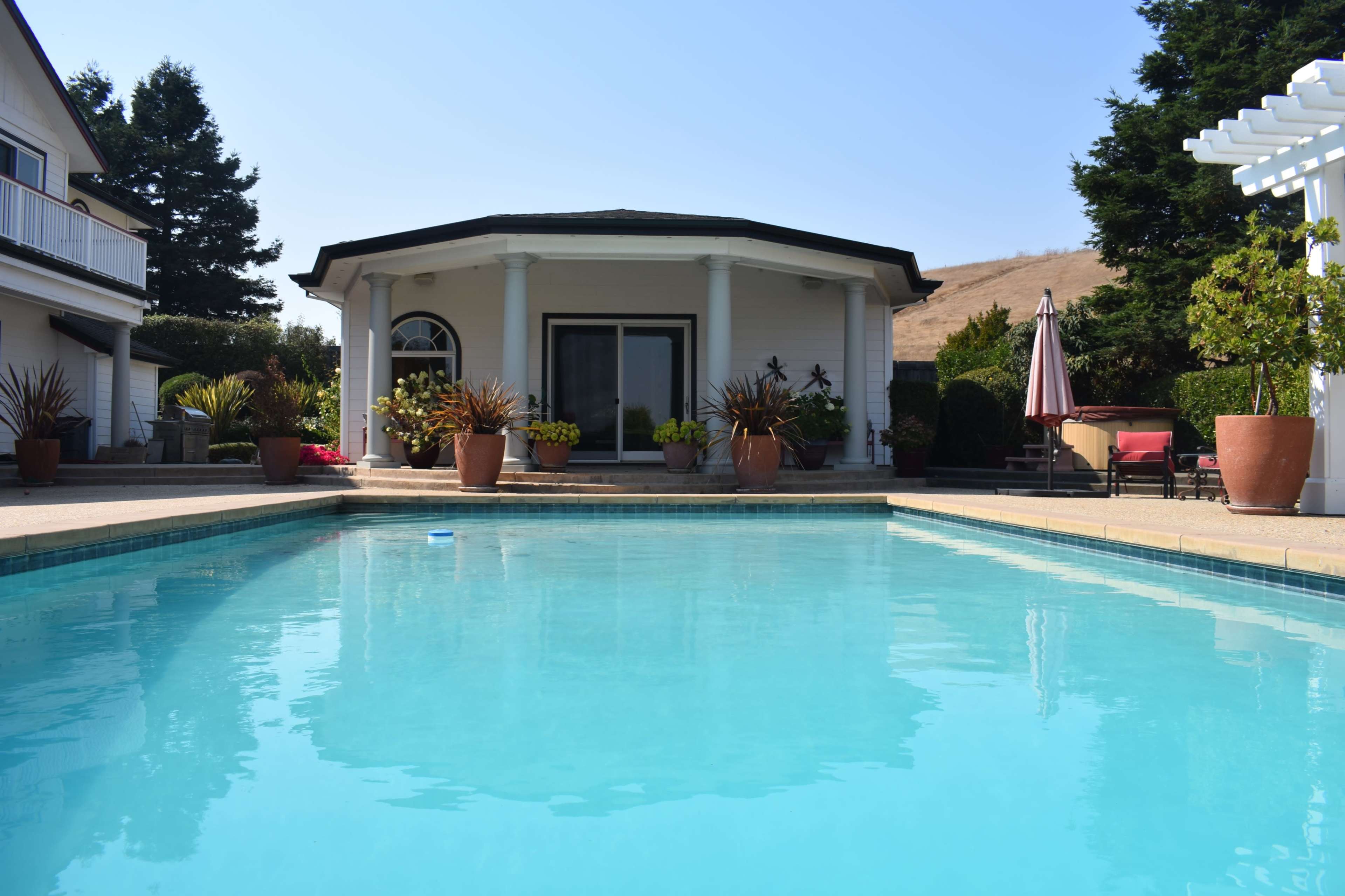 A clear blue swimming pool reflects the surrounding patio and a house with pillars and a circular roof.