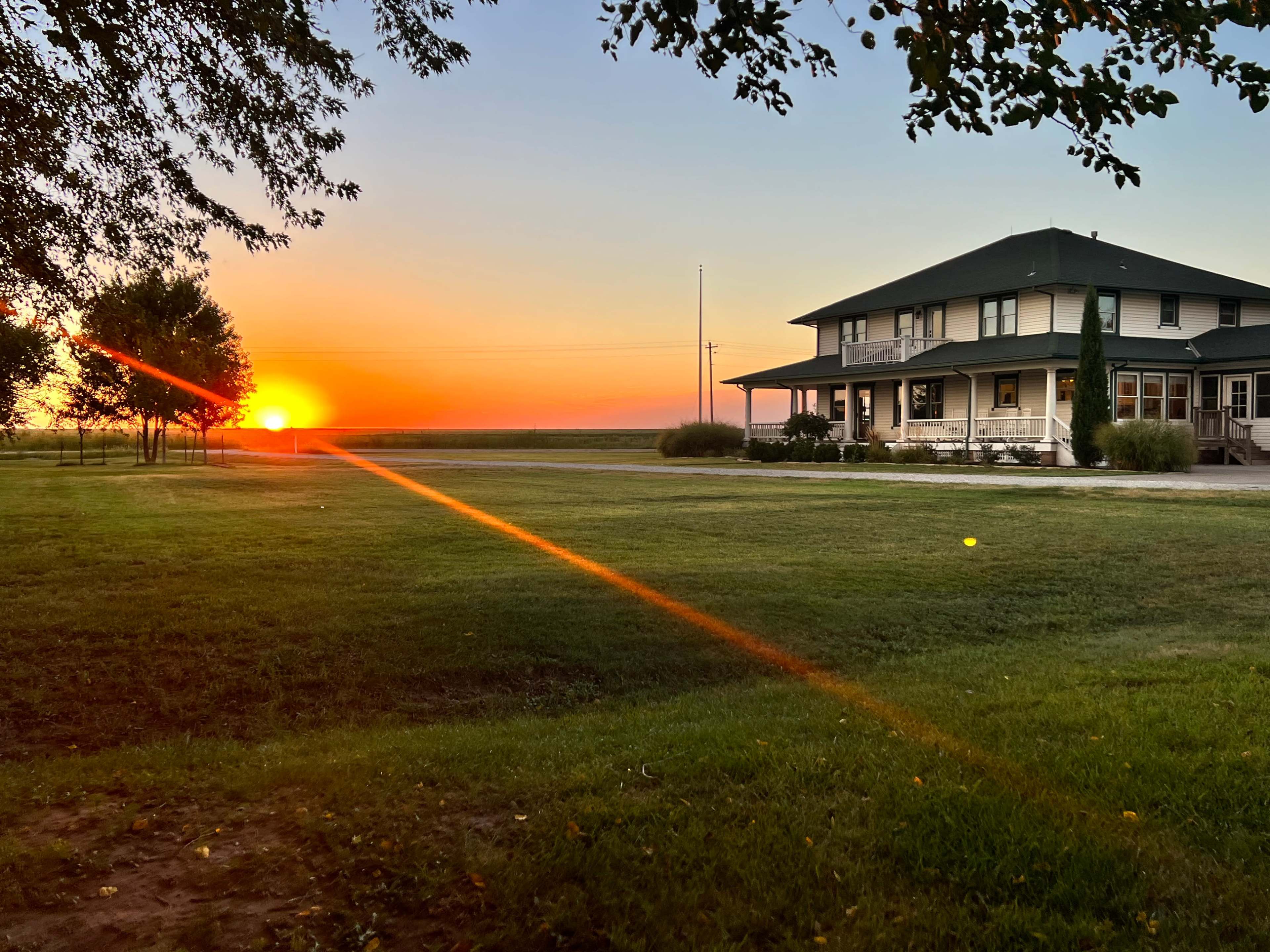 A large house with a wraparound porch sits on a grassy lawn, as the sun sets in the background, casting a warm glow over the scene.
