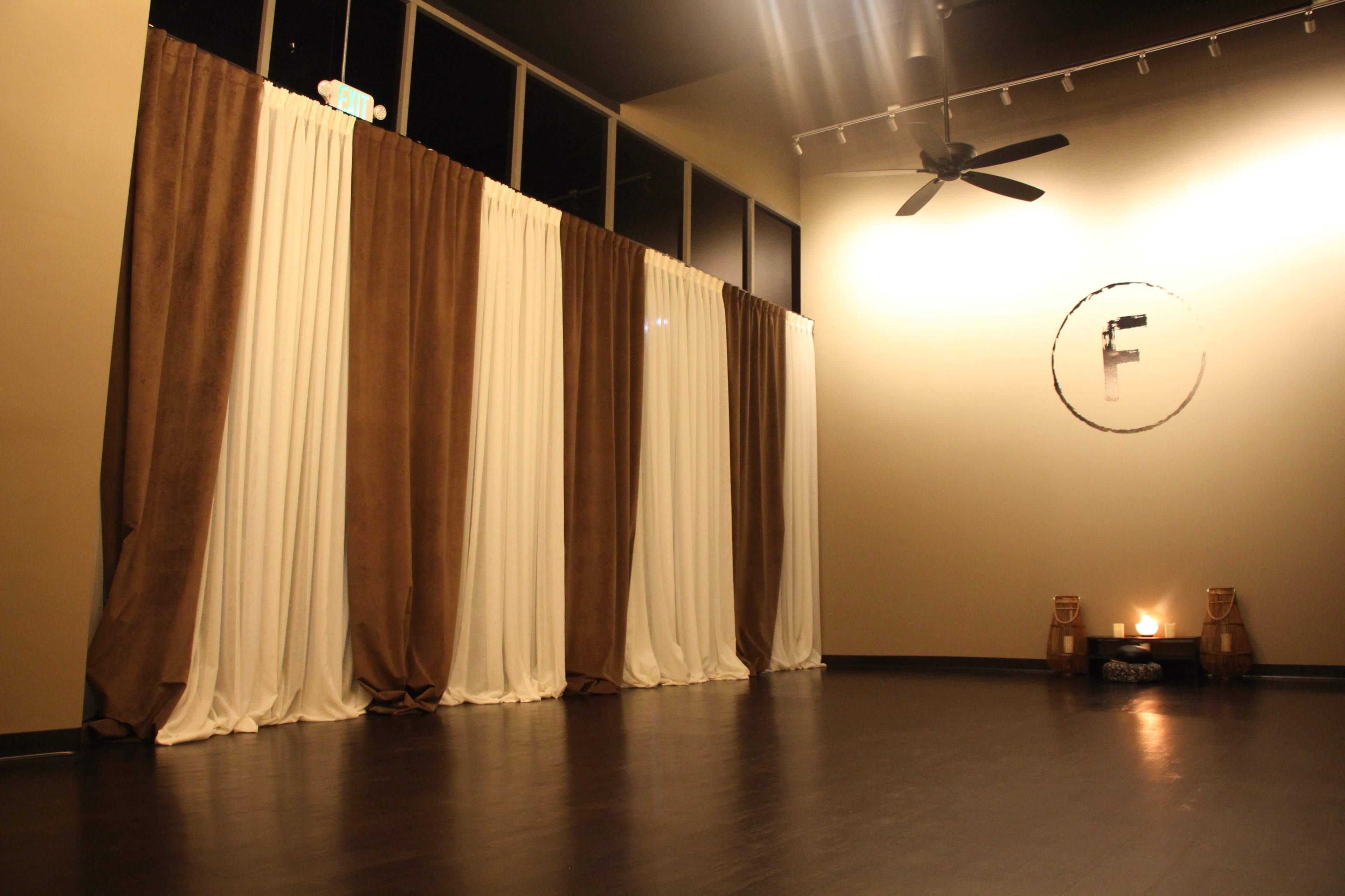 A spacious room with large, floor-to-ceiling curtains in brown and white, a fan on the ceiling, and a simple circular wall decoration with an "F" in the center, along with two candles on a table.